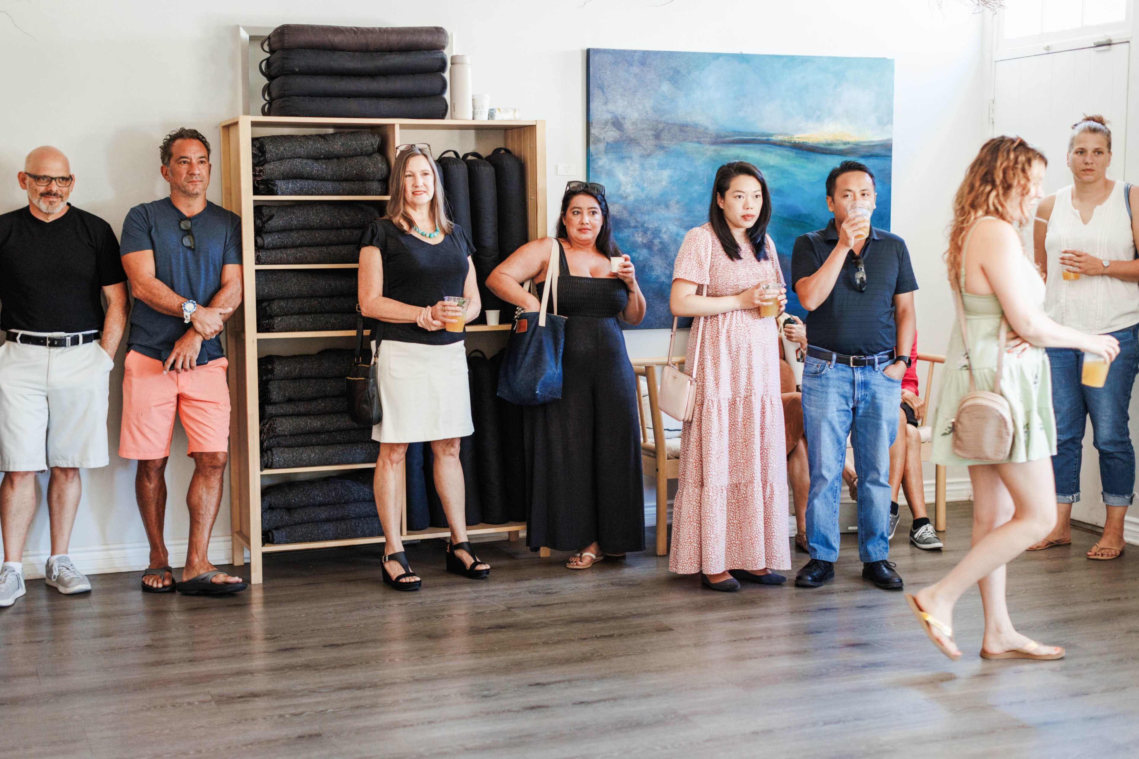 A group of people stands in a well-lit room with a shelf of folded blankets and a blue painting on the wall.