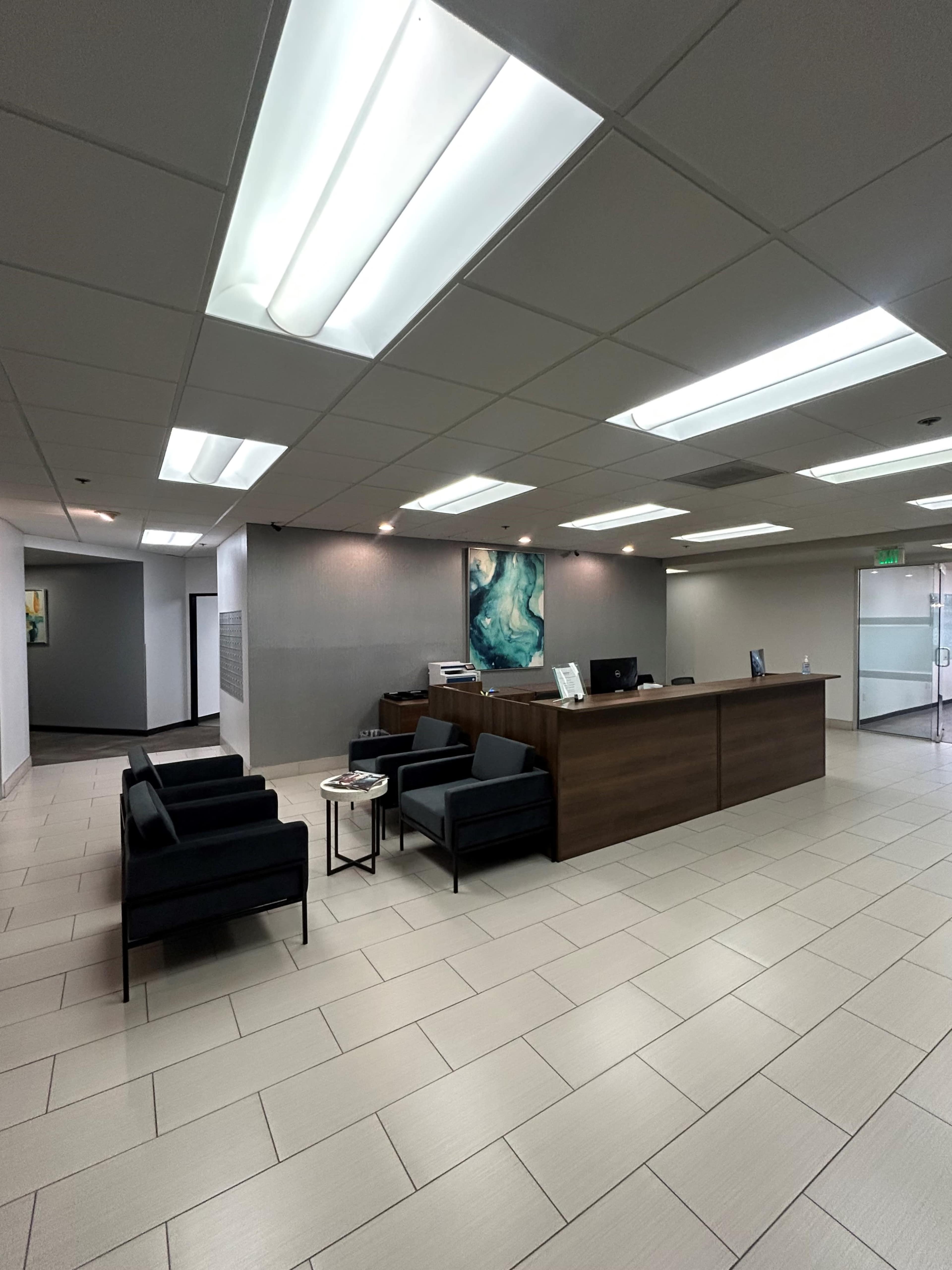 The image shows a modern reception area with a dark wood front desk, black chairs arranged for seating, and soft overhead lighting.