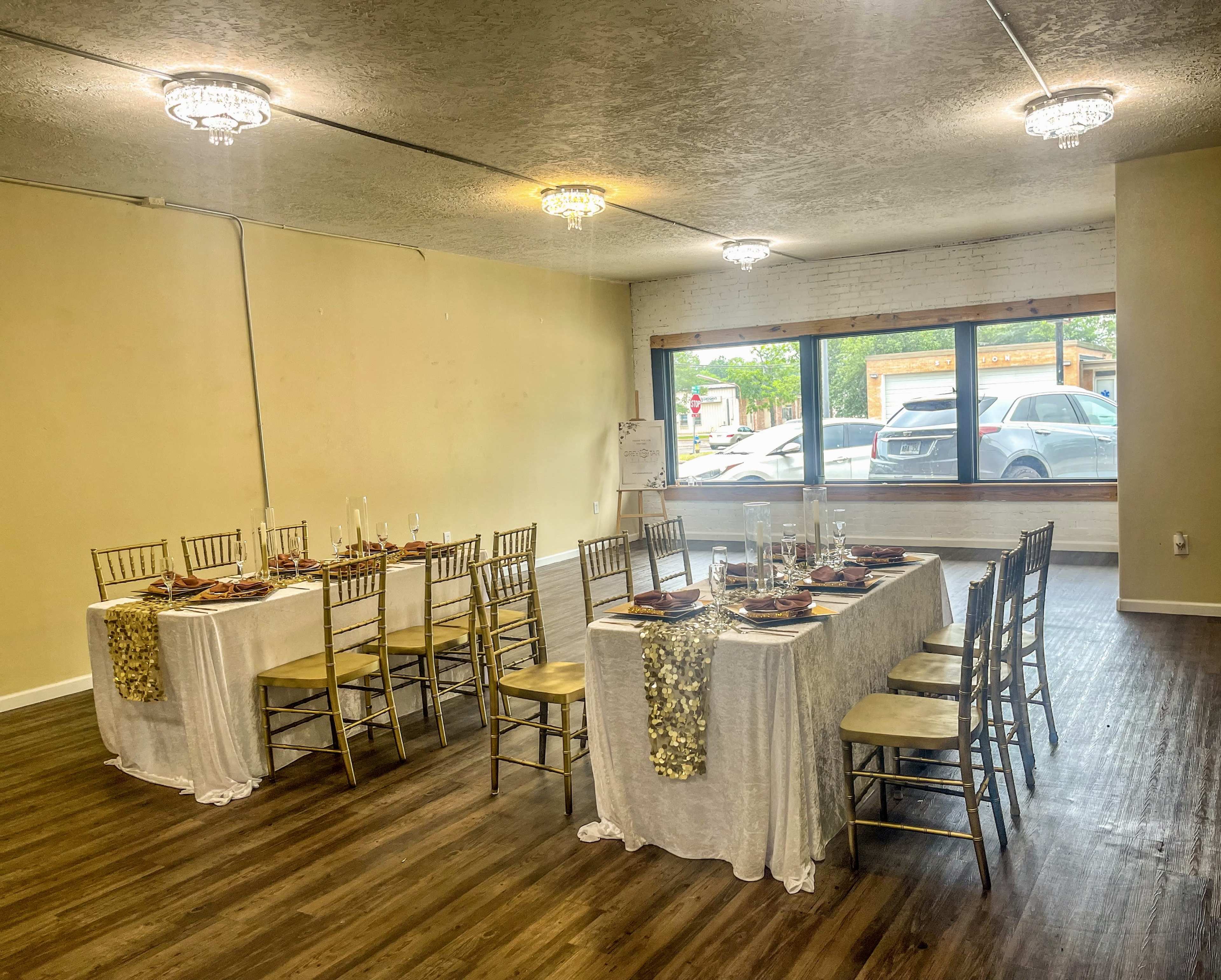 A long banquet table is set with dinnerware and candles in a well-lit room with large windows overlooking parked cars outside.