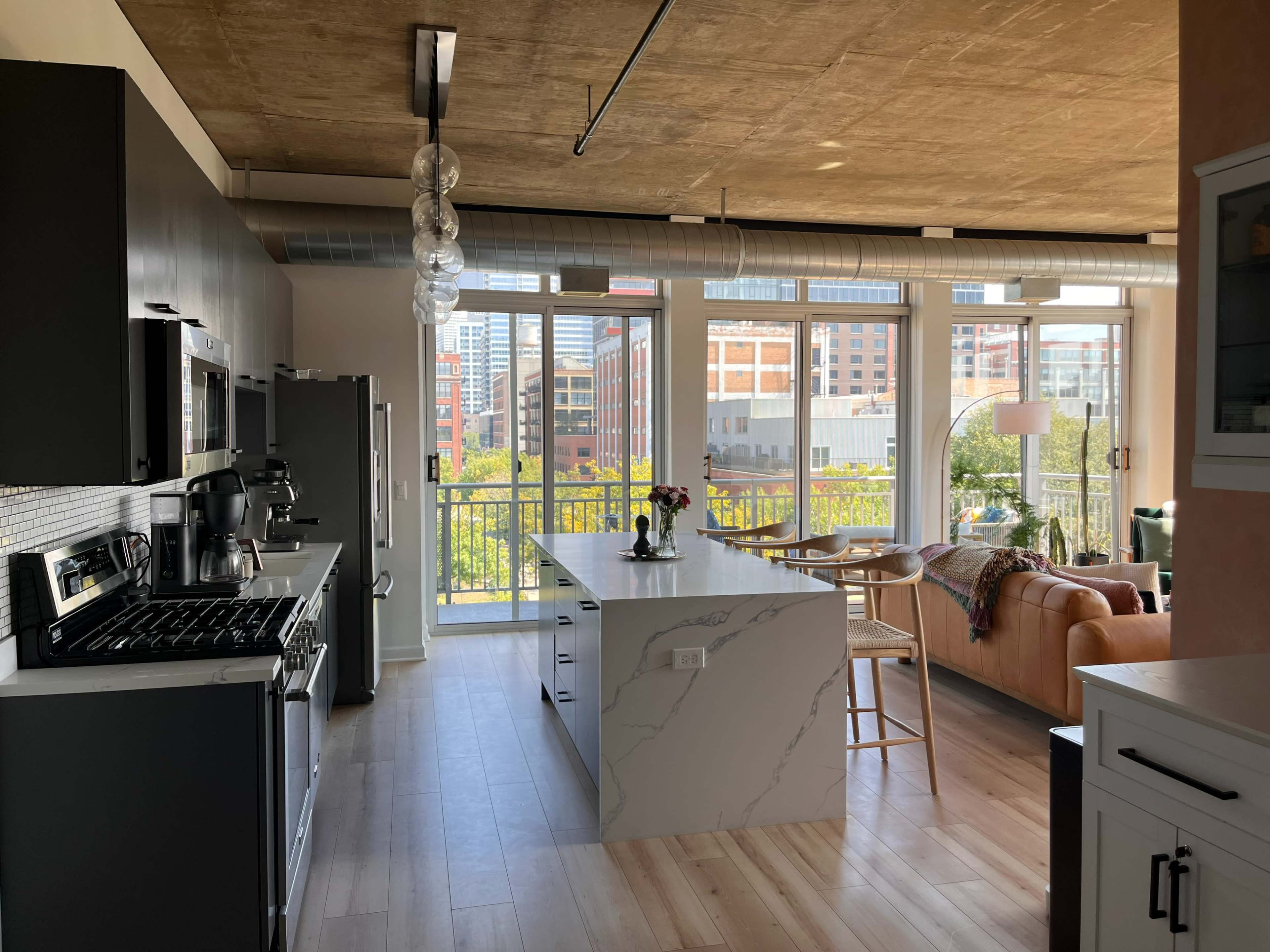 A modern kitchen and living area with a large island, stainless steel appliances, and a view of a city through large glass windows.