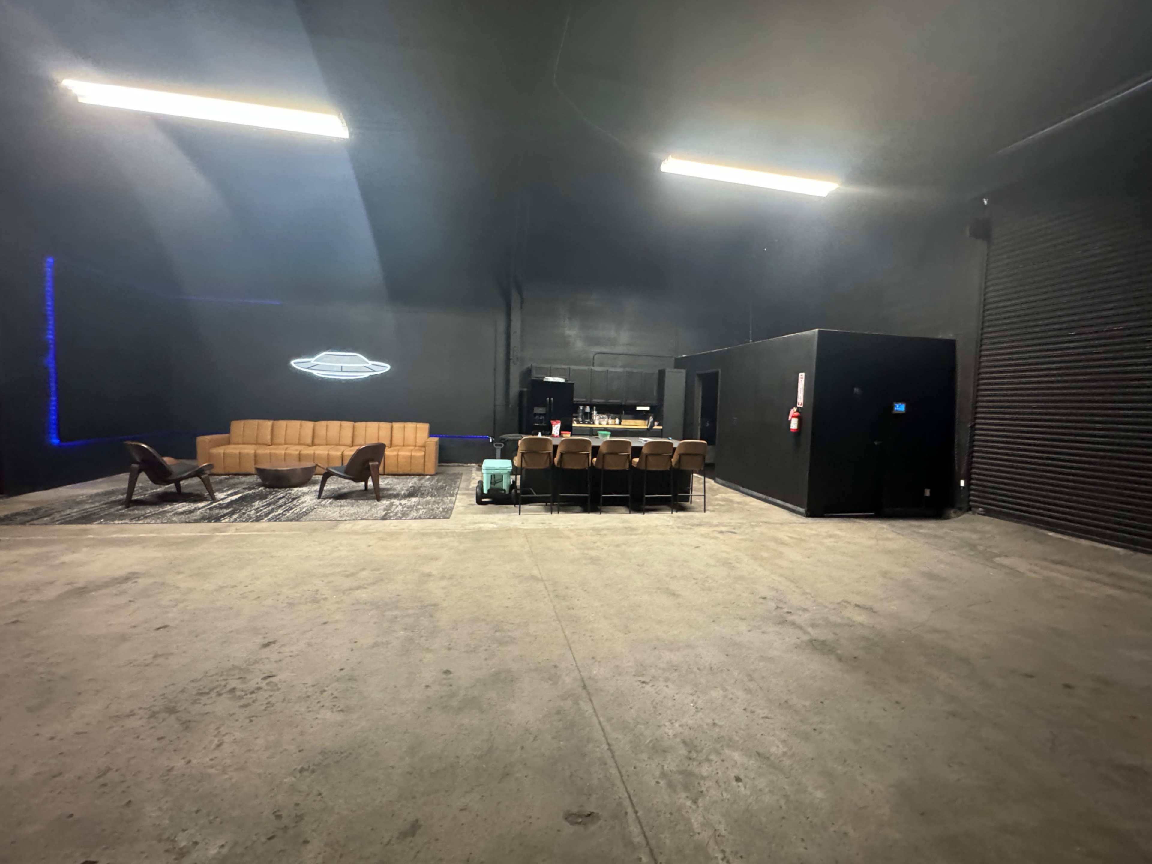 The image depicts a spacious, dimly lit indoor area featuring a modern seating arrangement with a couch, a bar setup, and a black storage unit against the far wall.