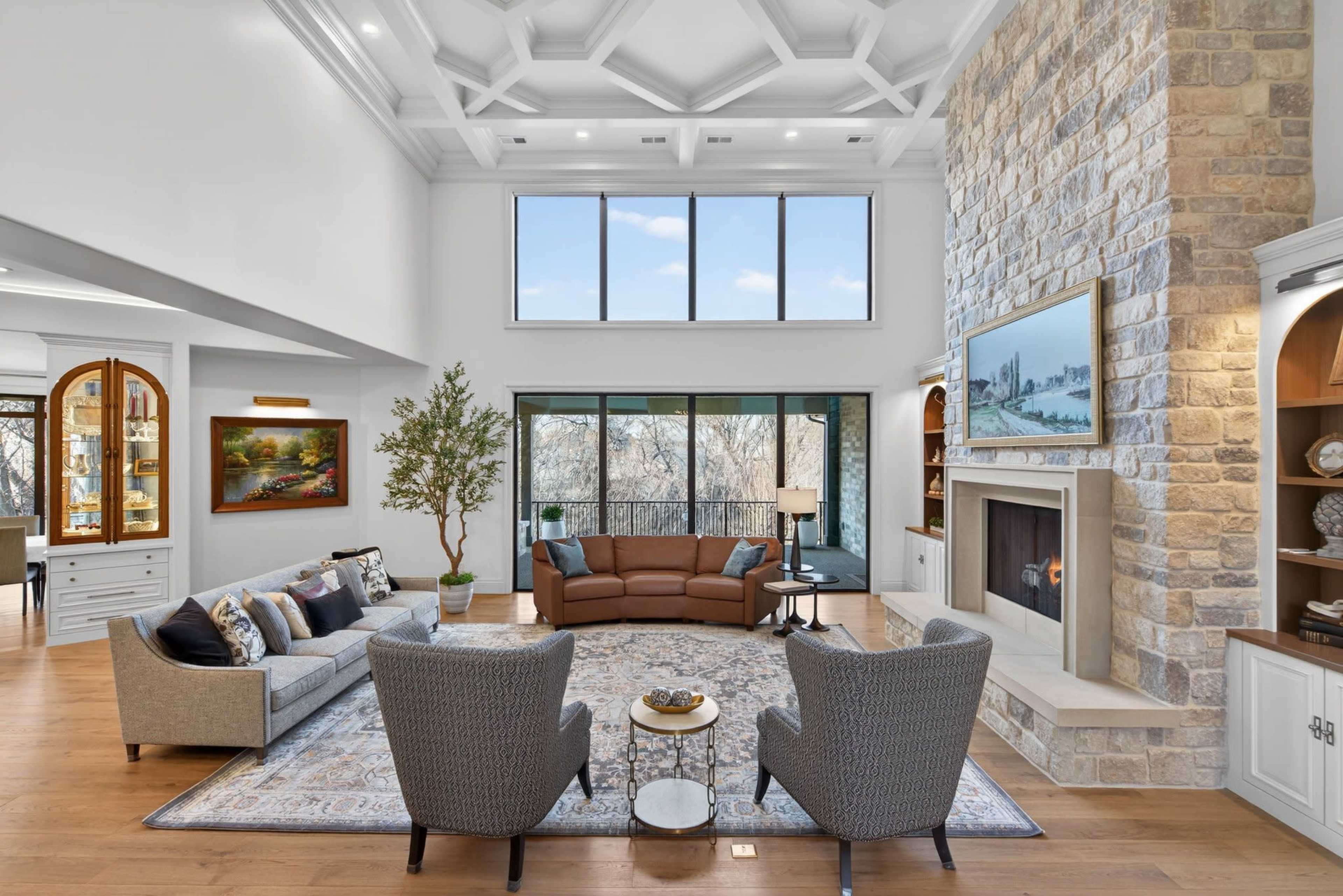 The image shows a spacious living room with a high ceiling, large windows, a stone fireplace, and a seating arrangement including a brown sofa and two gray armchairs.