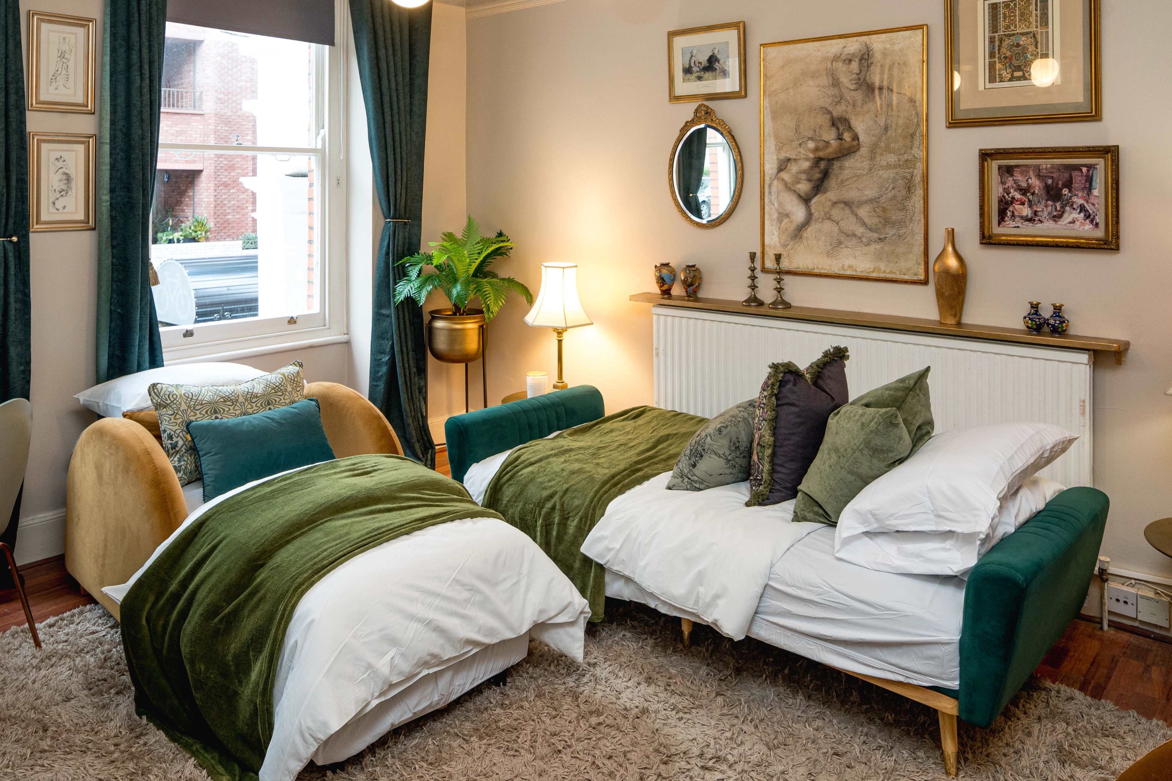 The image shows a cozy bedroom with two beds, one partially made, a large area rug, framed artwork on the walls, and decorative plants.