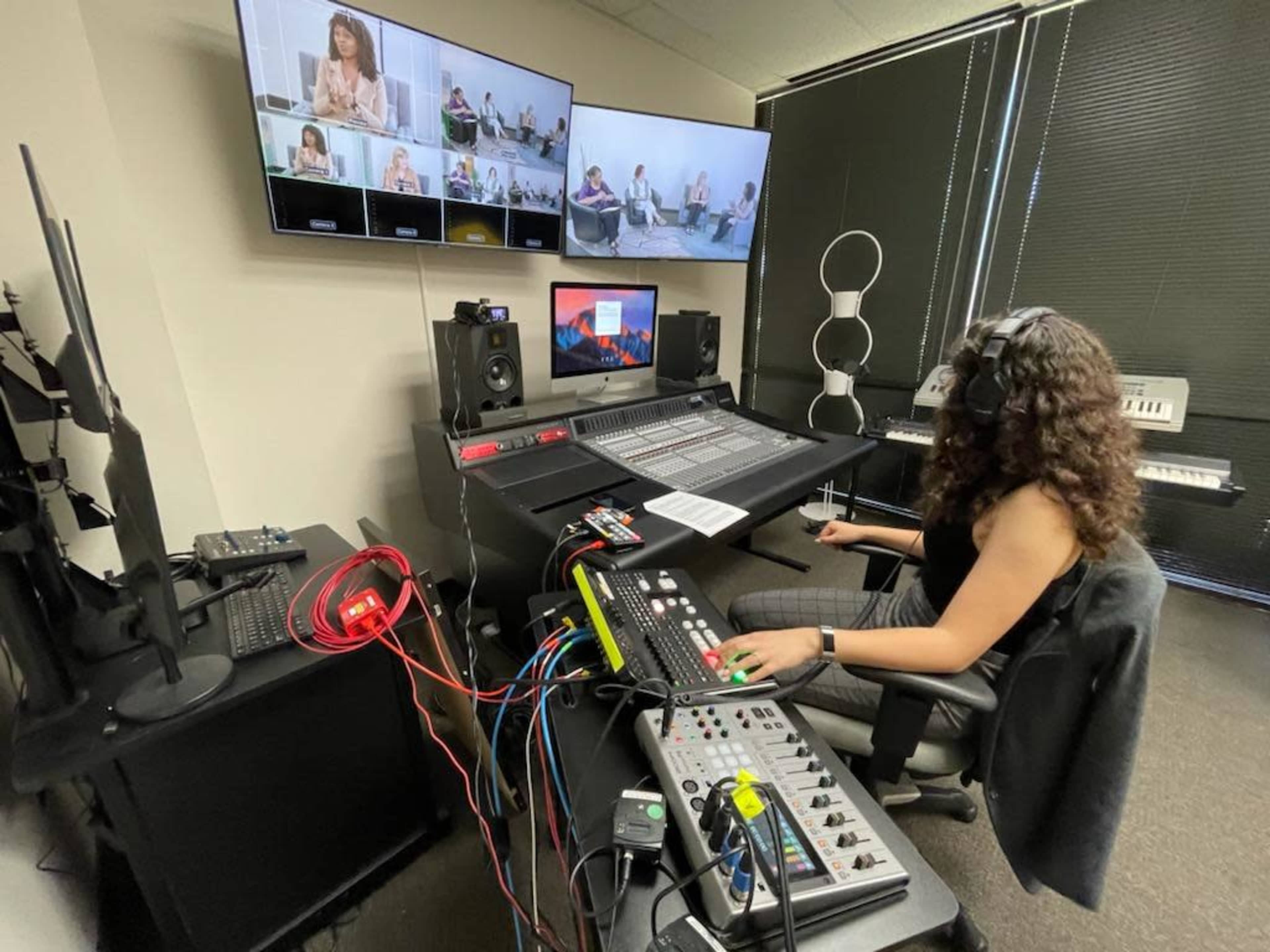 A person wearing headphones operates a sound mixing board in a control room with multiple screens displaying virtual participants.