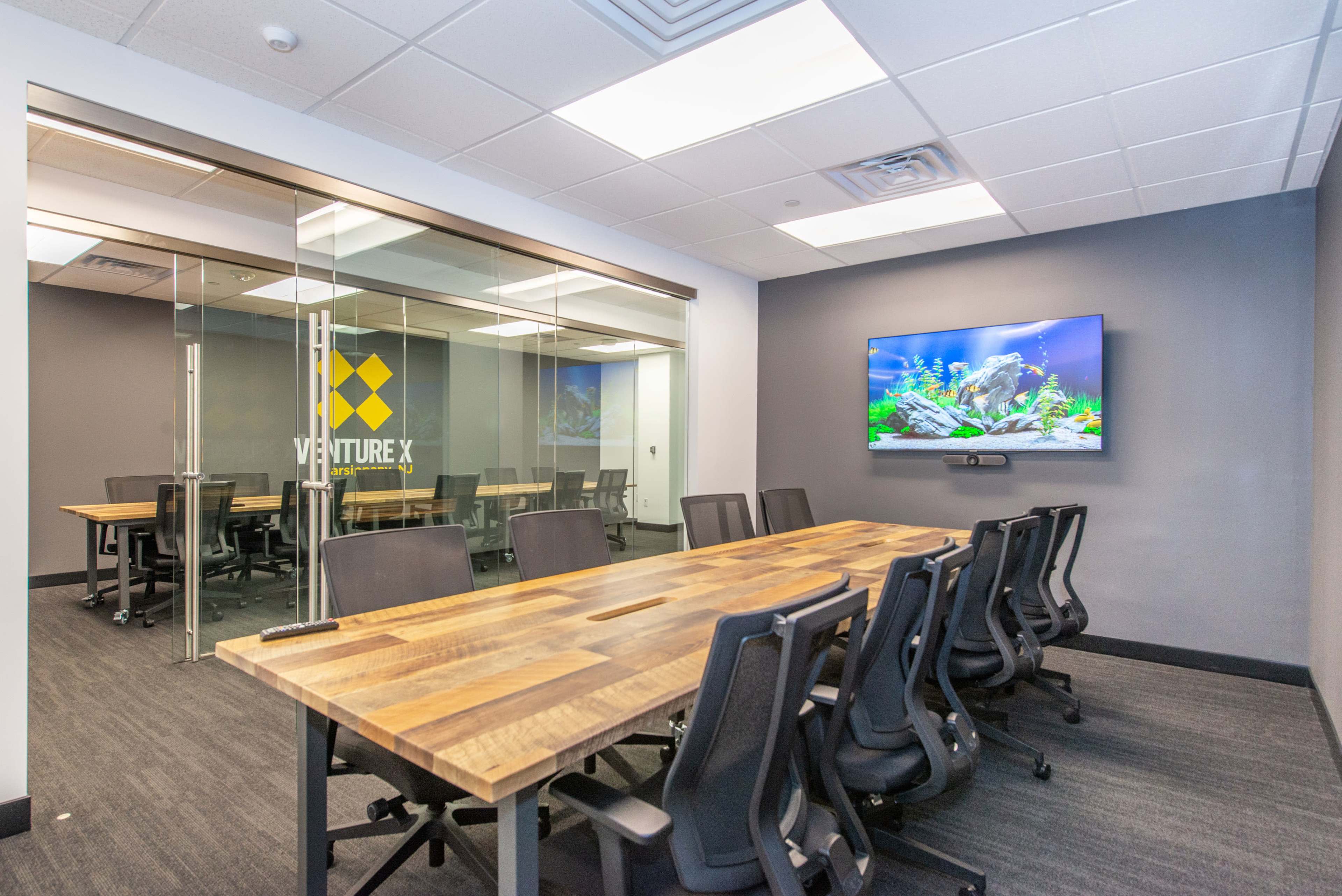 A modern conference room features a wooden table with black ergonomic chairs, a wall-mounted television, and a view of an aquarium.