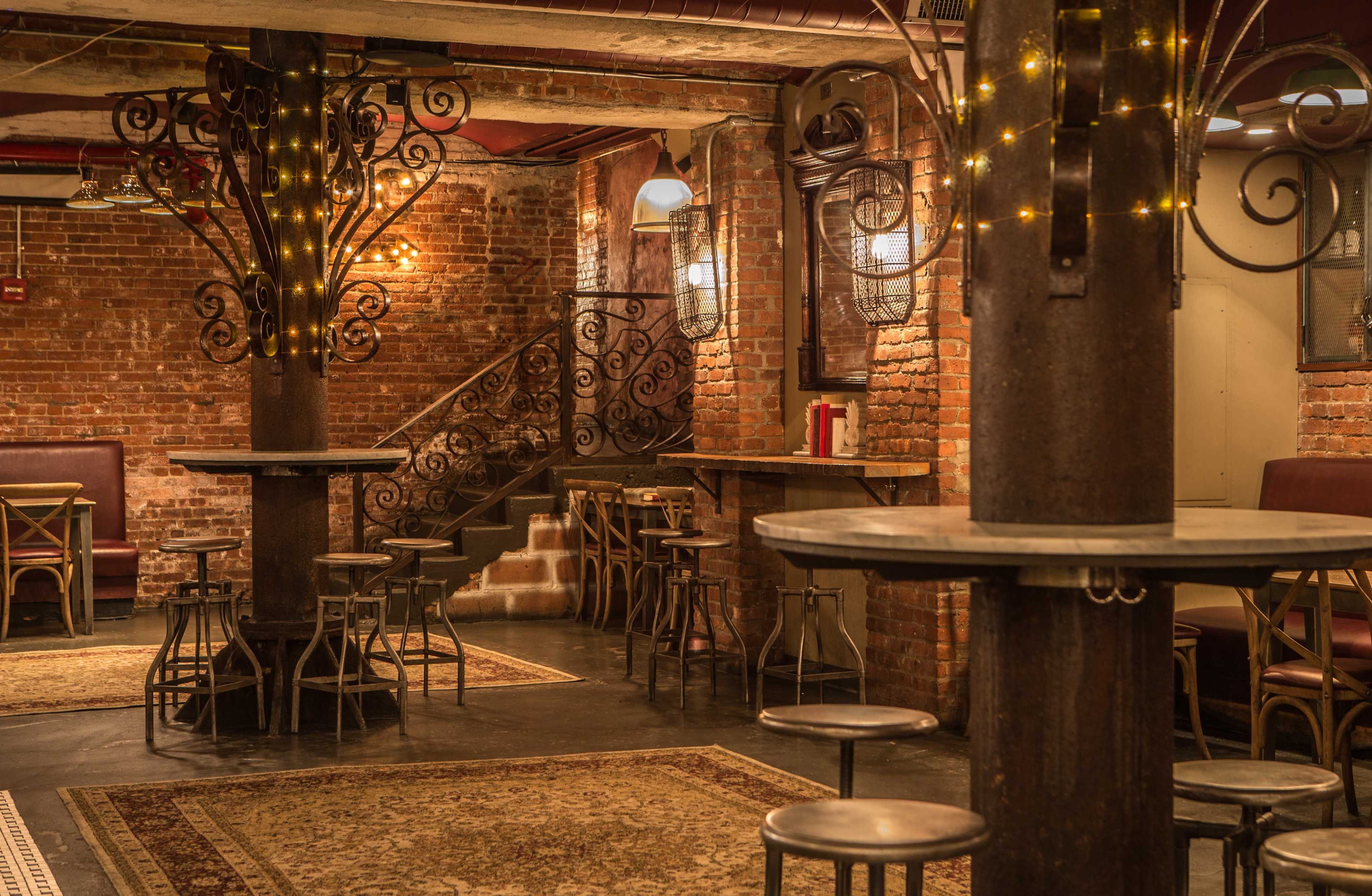 The image shows an intimate bar area with brick walls, wrought iron details, and wooden tables surrounded by metal stools.