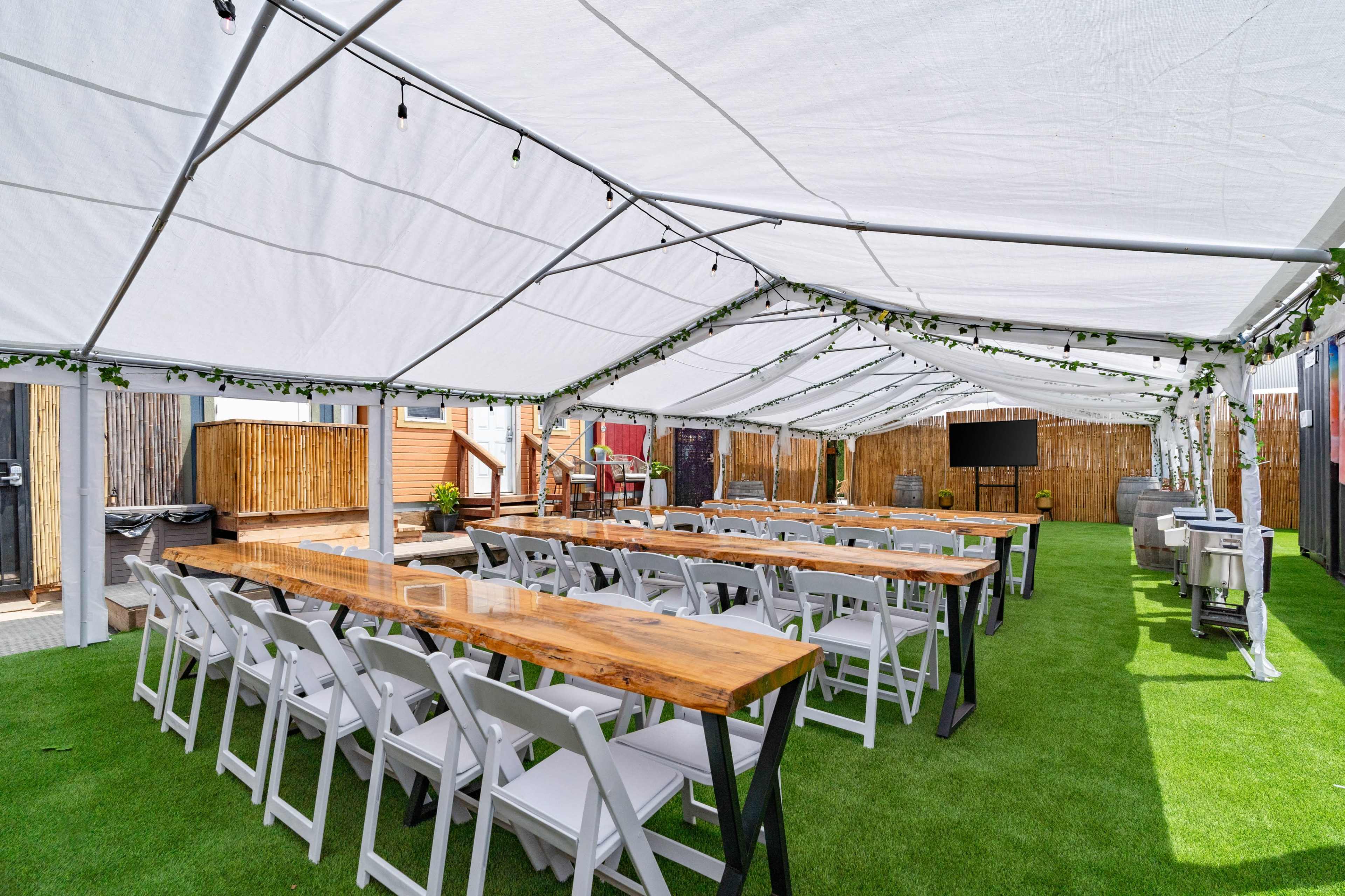 A large outdoor tent set up for an event features long wooden tables and white chairs arranged on green turf.