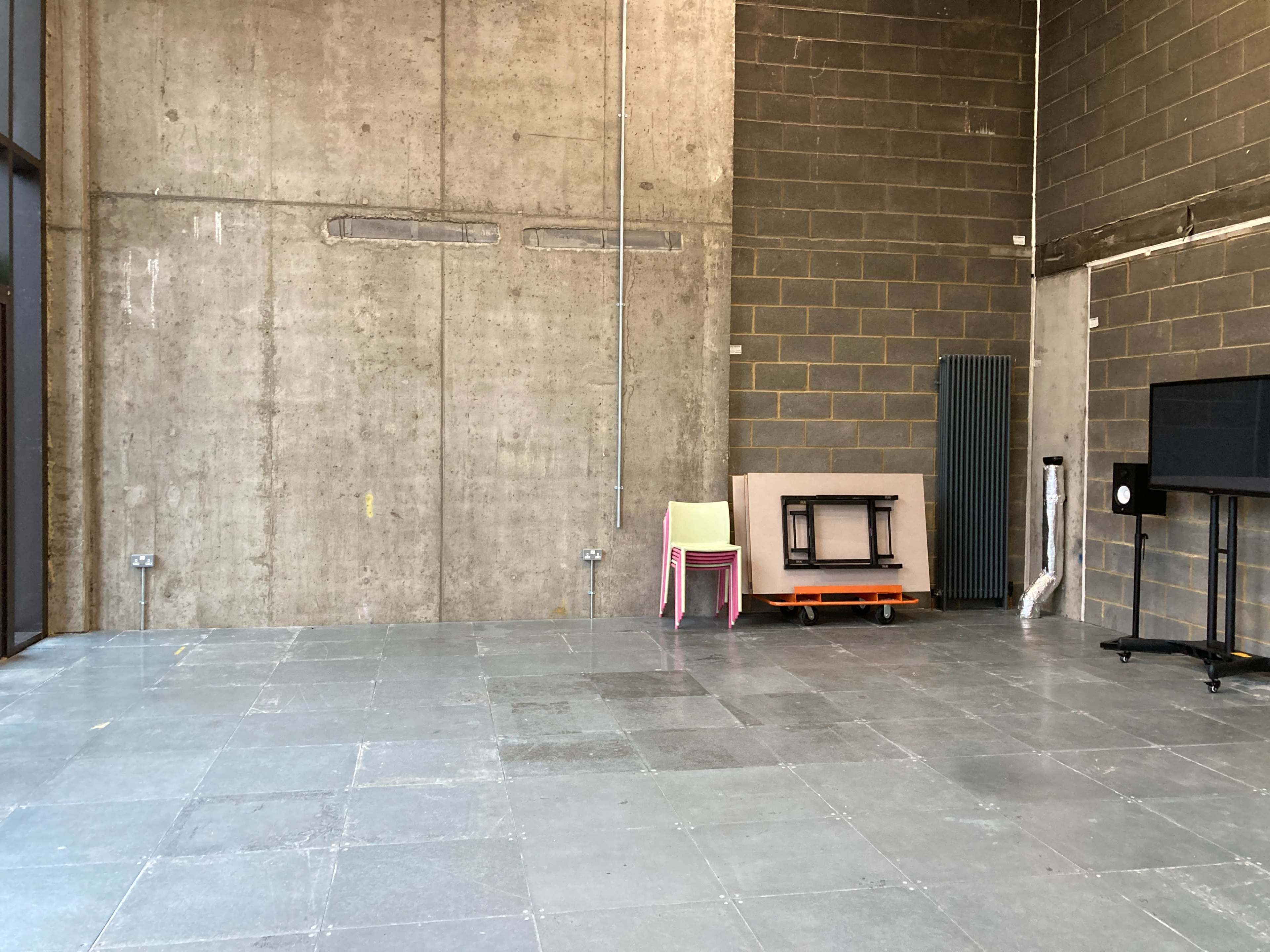An empty industrial space with a concrete wall, gray tiled floor, and a few pieces of furniture, including stacked chairs and a black television stand.
