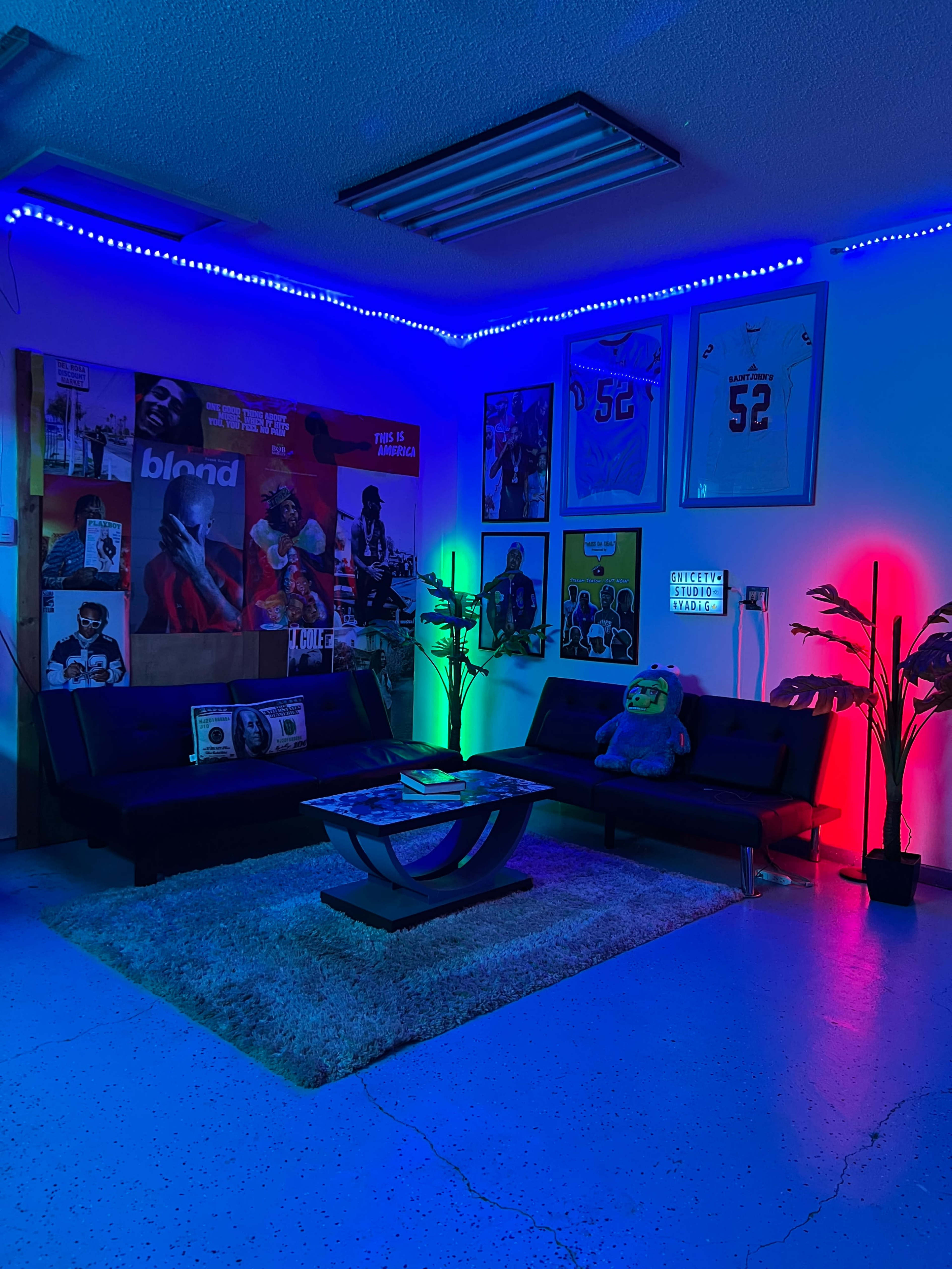 The image shows a modern lounge area featuring a black sofa set, a coffee table, and colorful ambient lighting with various framed artworks and posters on the walls.