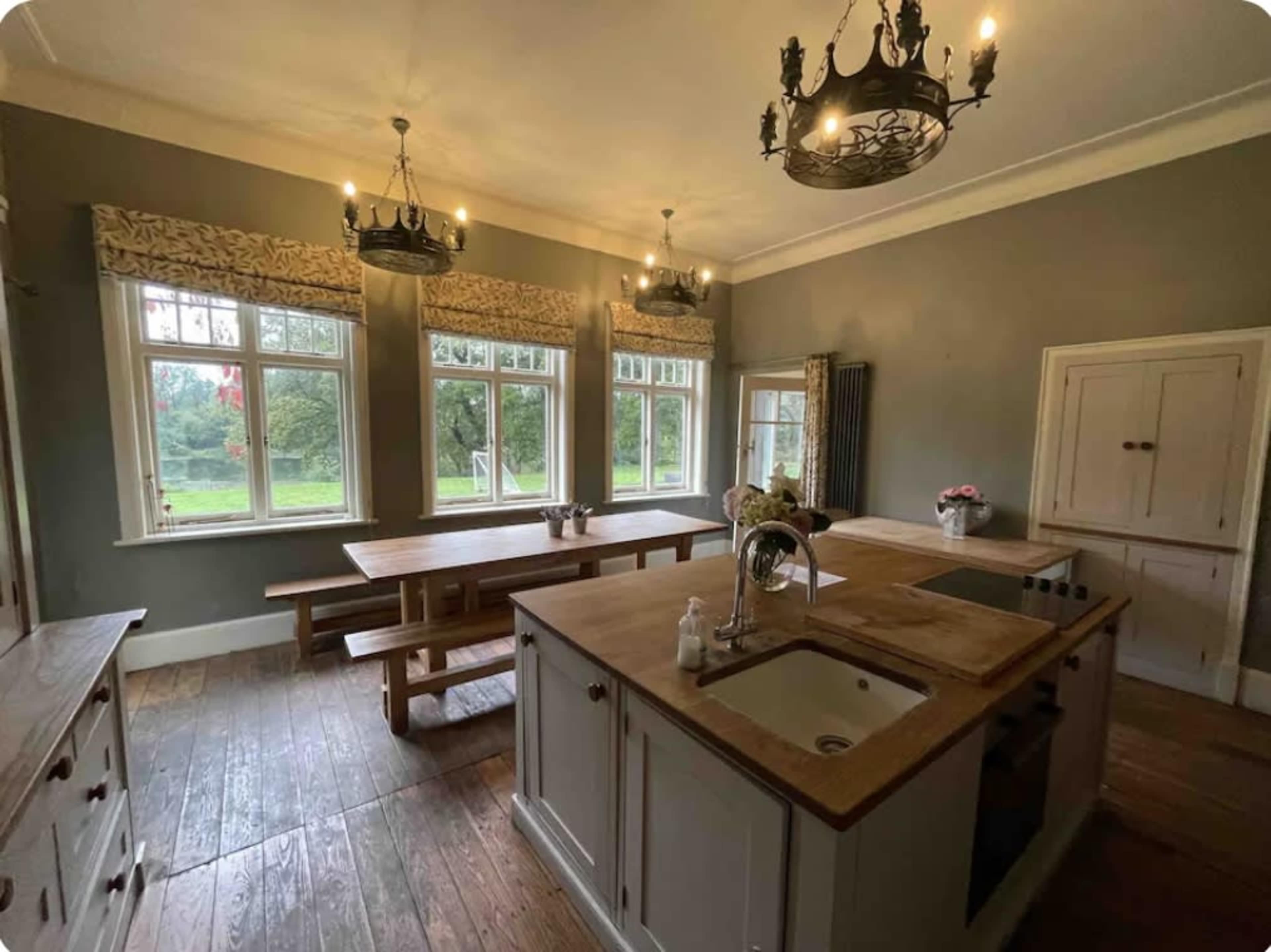 The kitchen features a central island with a sink, wooden countertops, and a large dining table, illuminated by several chandeliers and natural light from the windows.