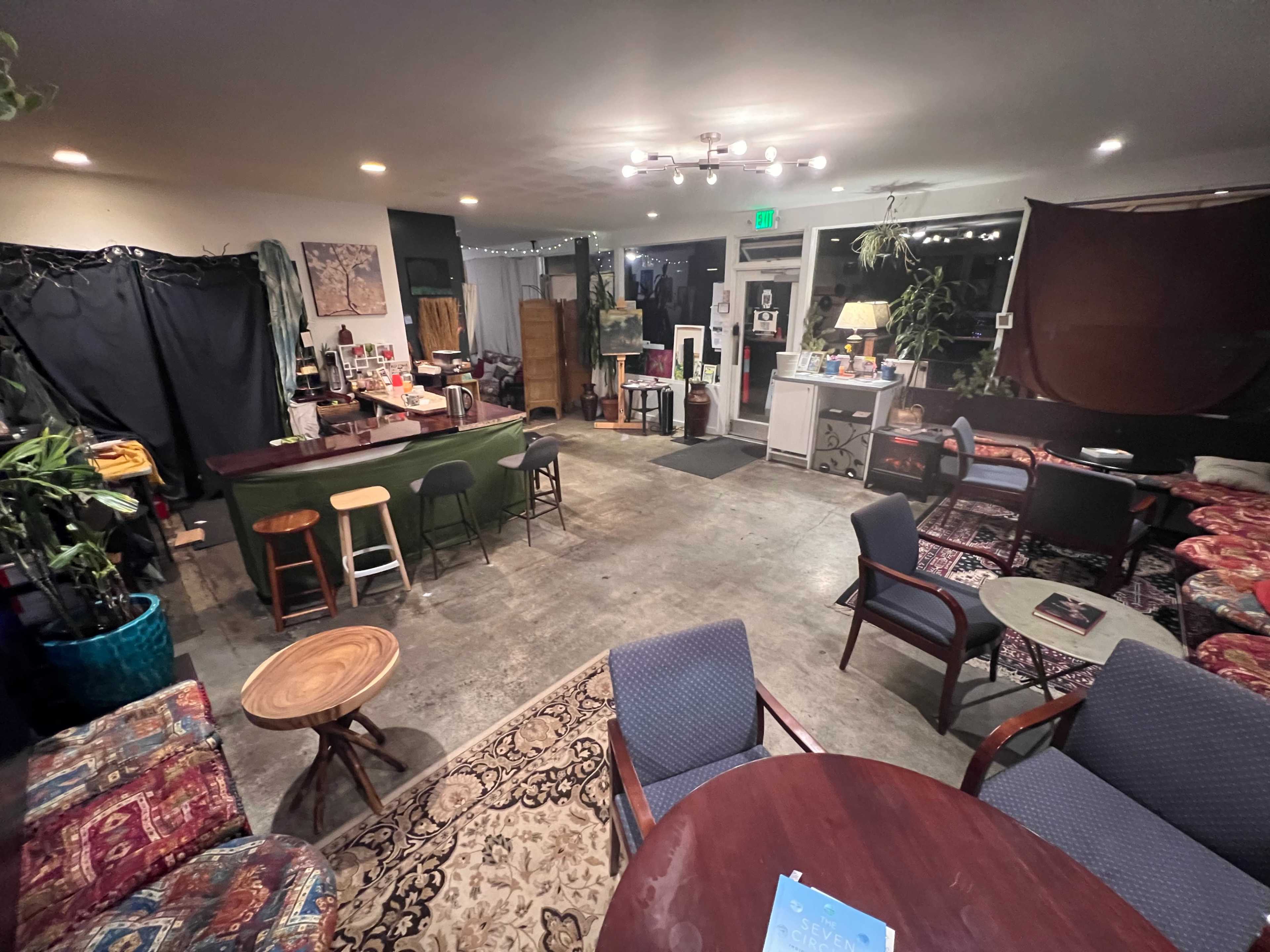 The image shows a cozy, dimly lit café with various seating arrangements, including chairs and small tables, and a bar area in the background.