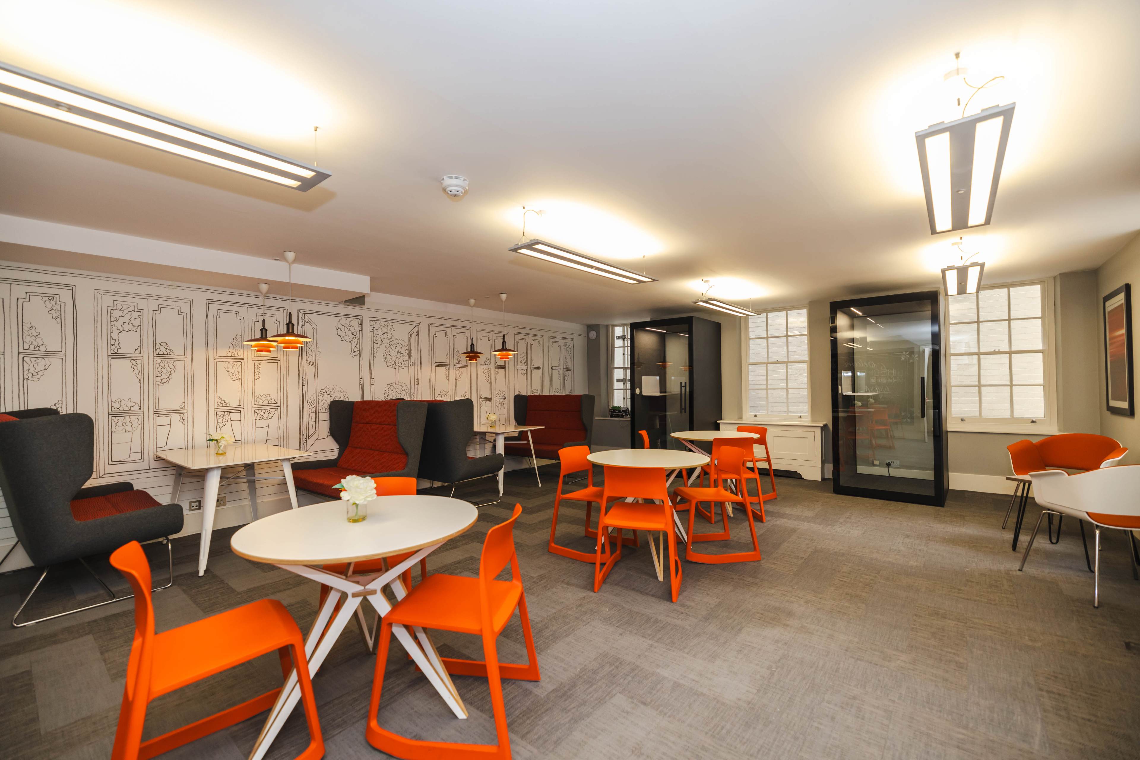 A modern lounge area with orange chairs and tables, featuring a gray carpet and decorative wall panels.