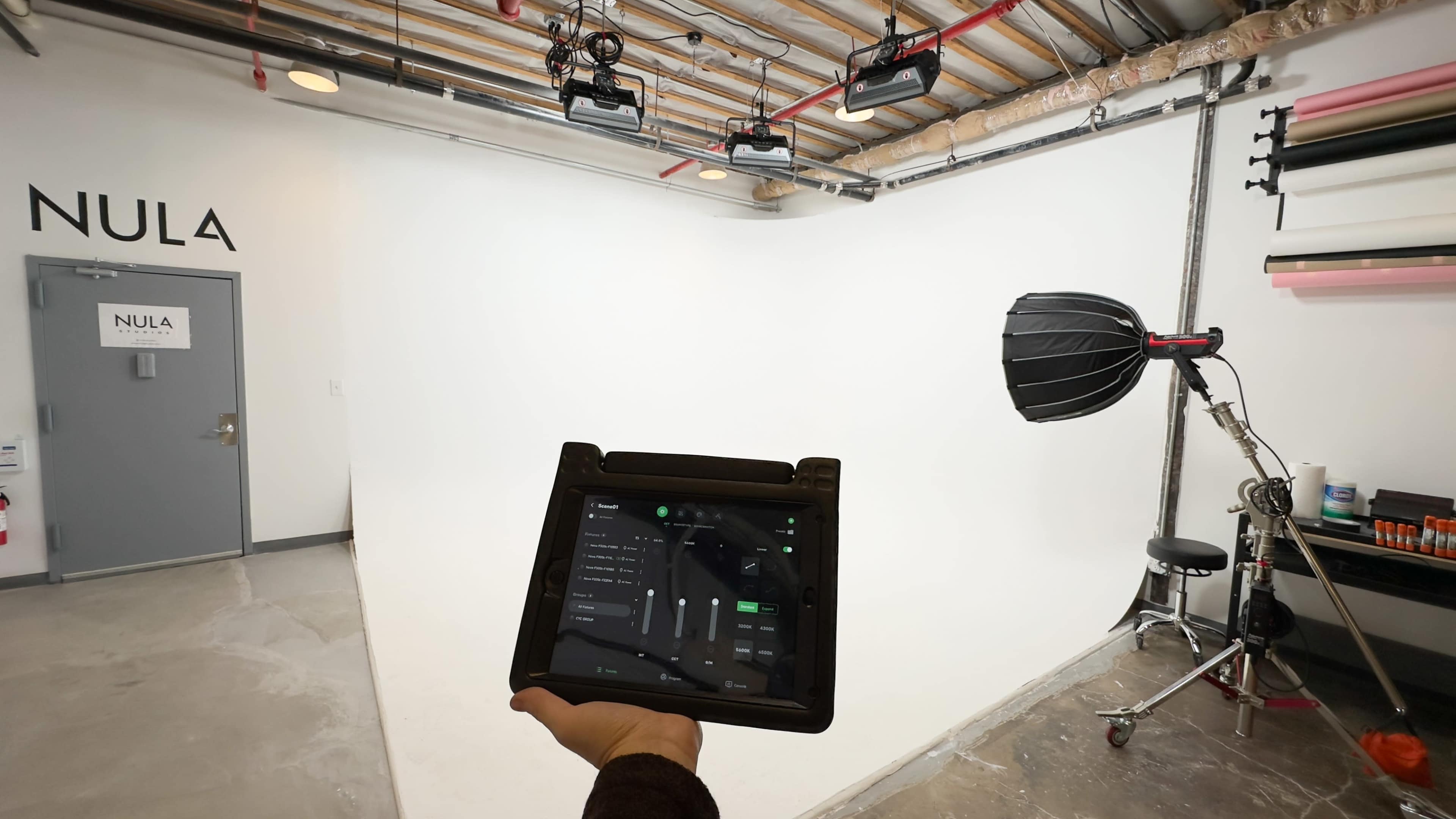 A person holds a control tablet in front of a photography studio with a white backdrop and lighting equipment.