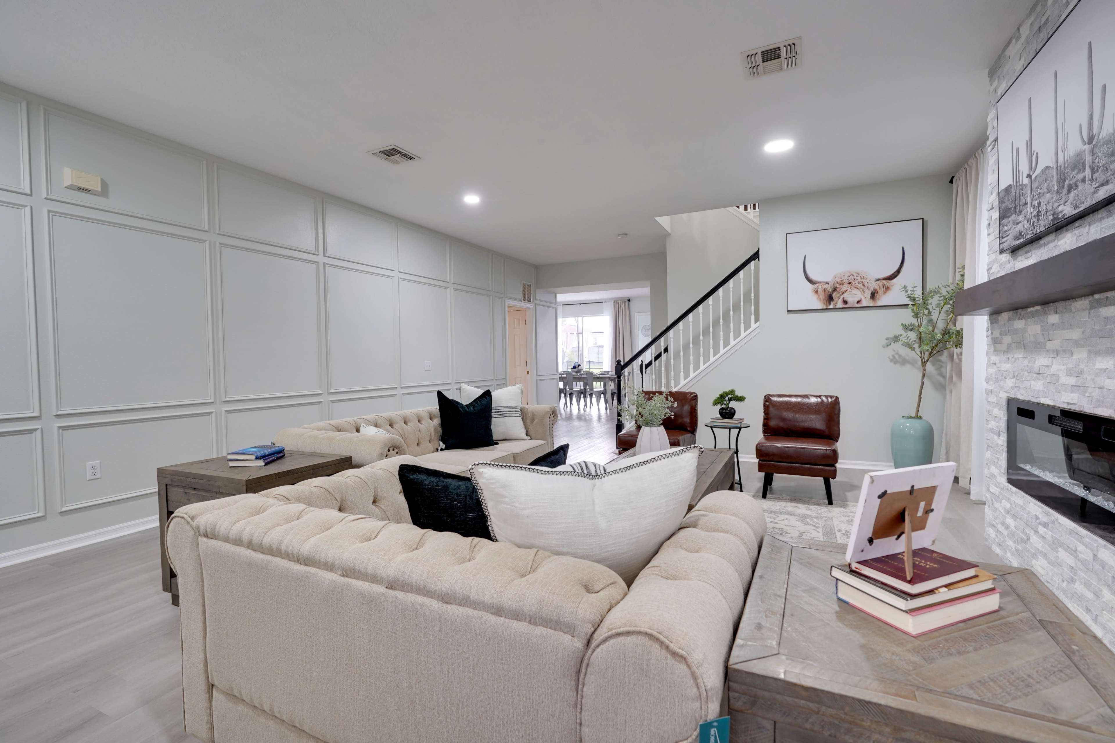 A modern living room with a light-colored tufted sofa, a coffee table with books, and a decorative plant, leading to a staircase and a dining area in the background.