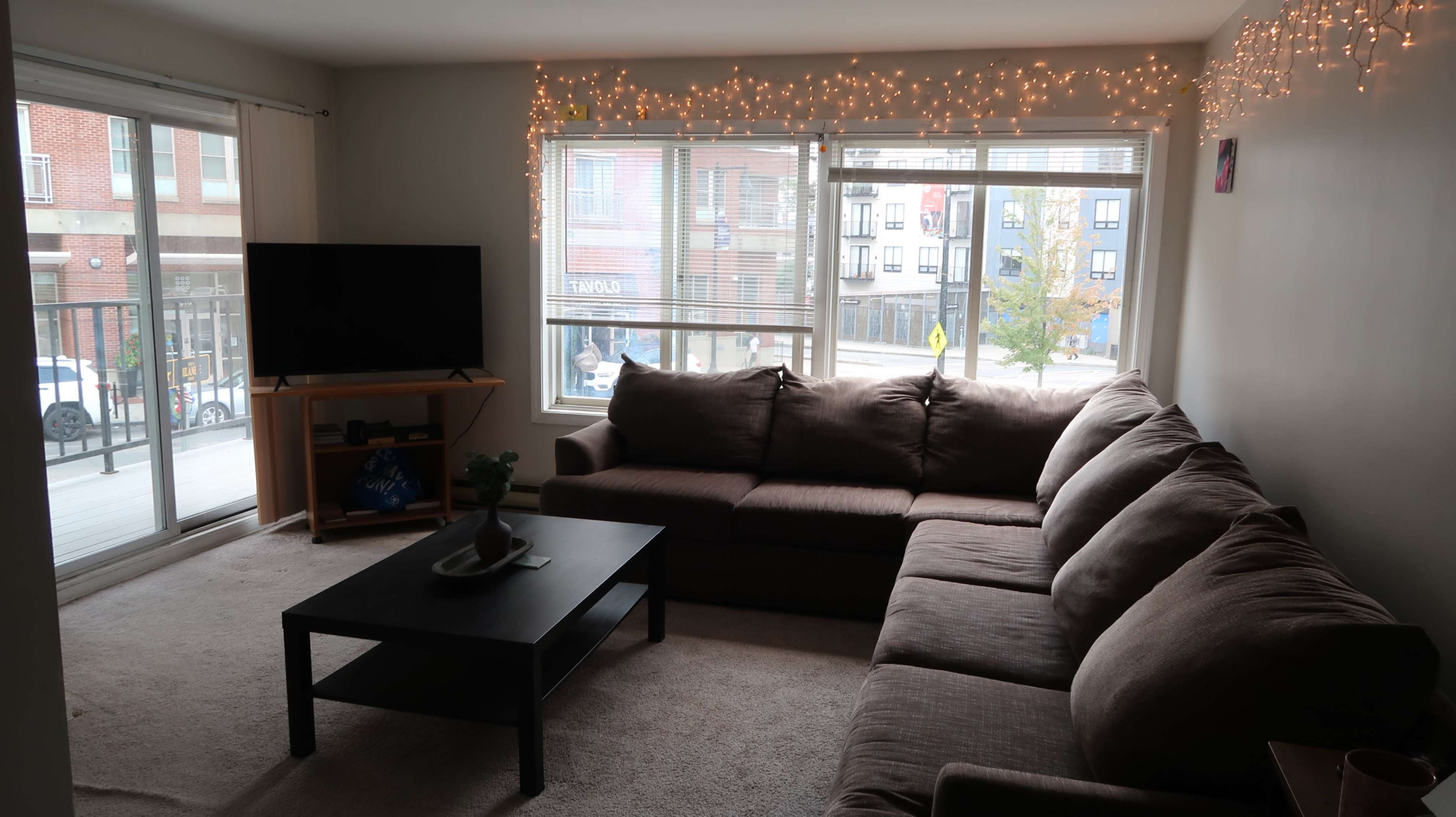 The image shows a living room with a large brown sectional sofa, a black coffee table, a television on a stand, and window views of an urban street decorated with string lights.