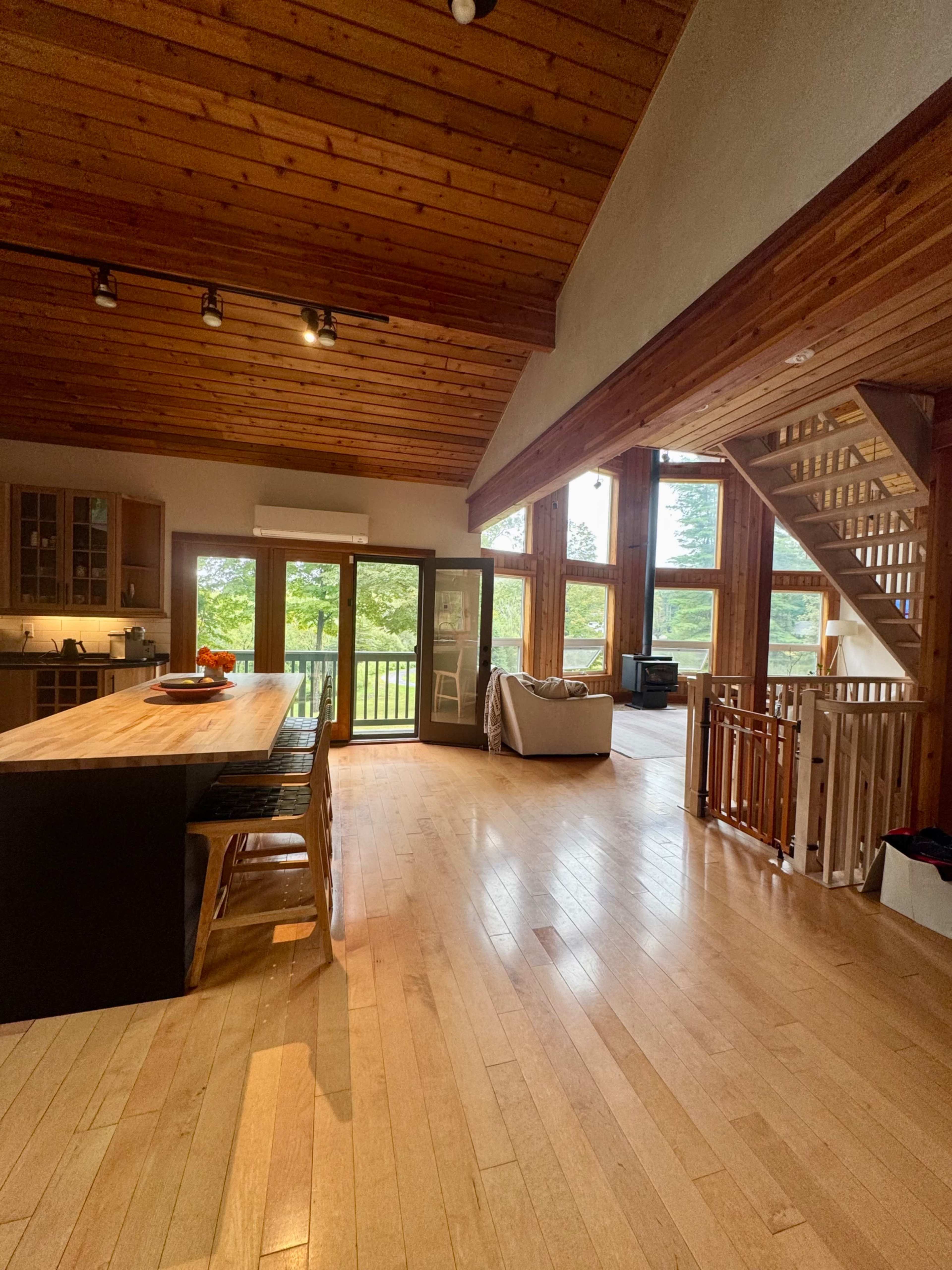 The image shows a spacious interior of a wooden-themed living area with a large table, kitchen area, and large windows allowing natural light to enter.