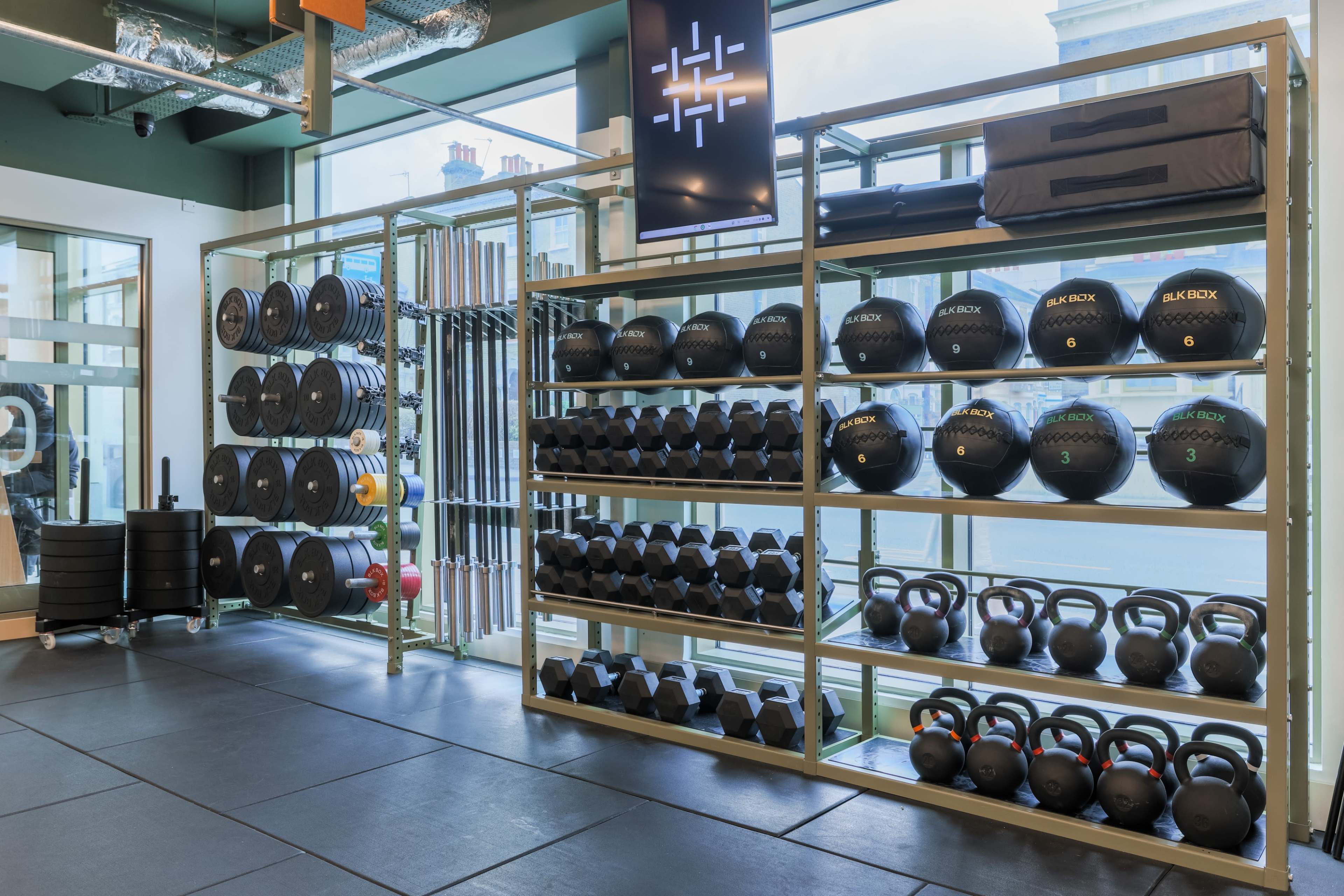 A fitness facility with organized racks of free weights, including dumbbells, kettlebells, and weight plates, against a backdrop of large windows.