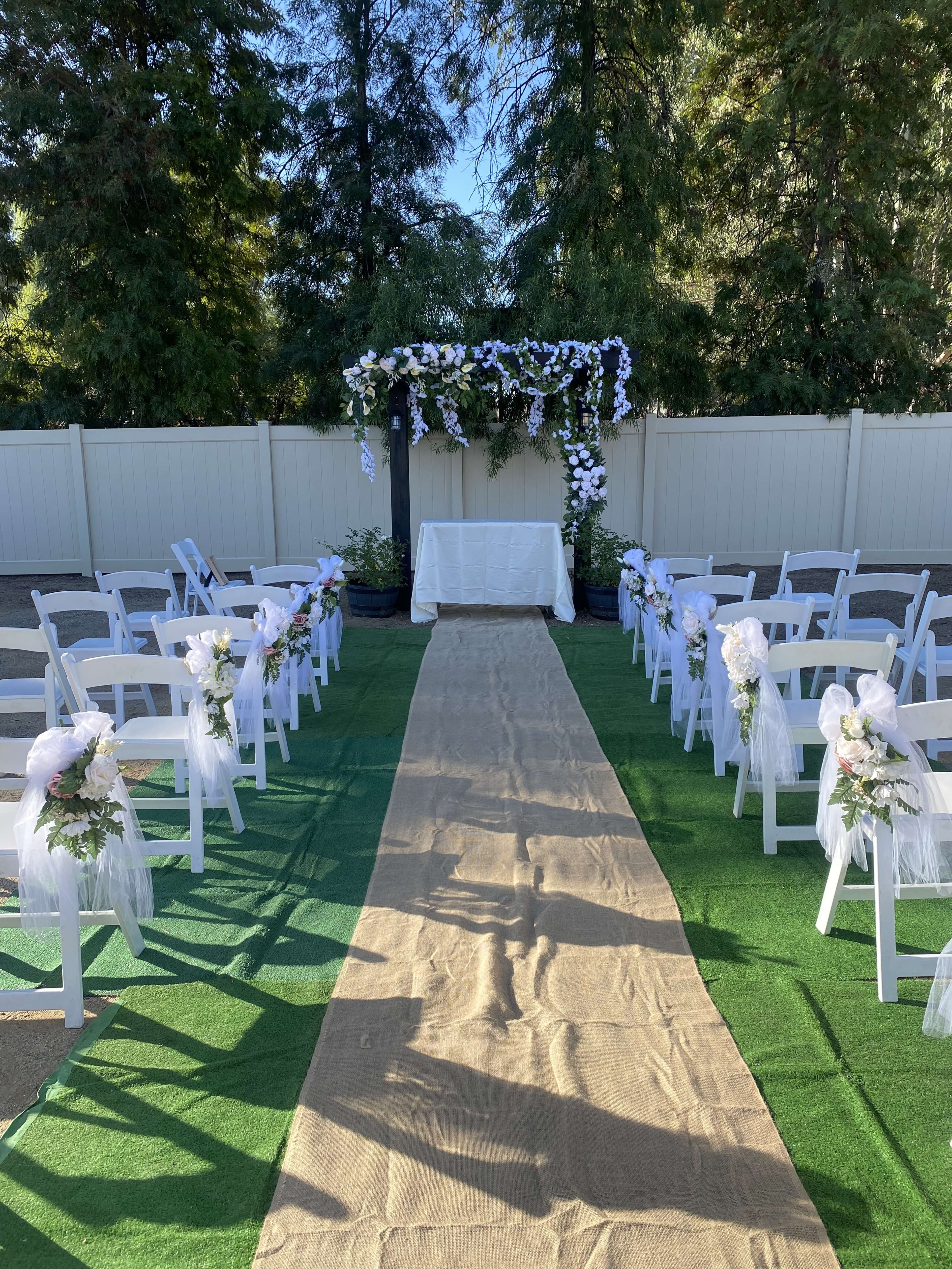 An outdoor wedding setup features white chairs arranged in rows along a central burlap aisle leading to a decorated altar with an arch of flowers.