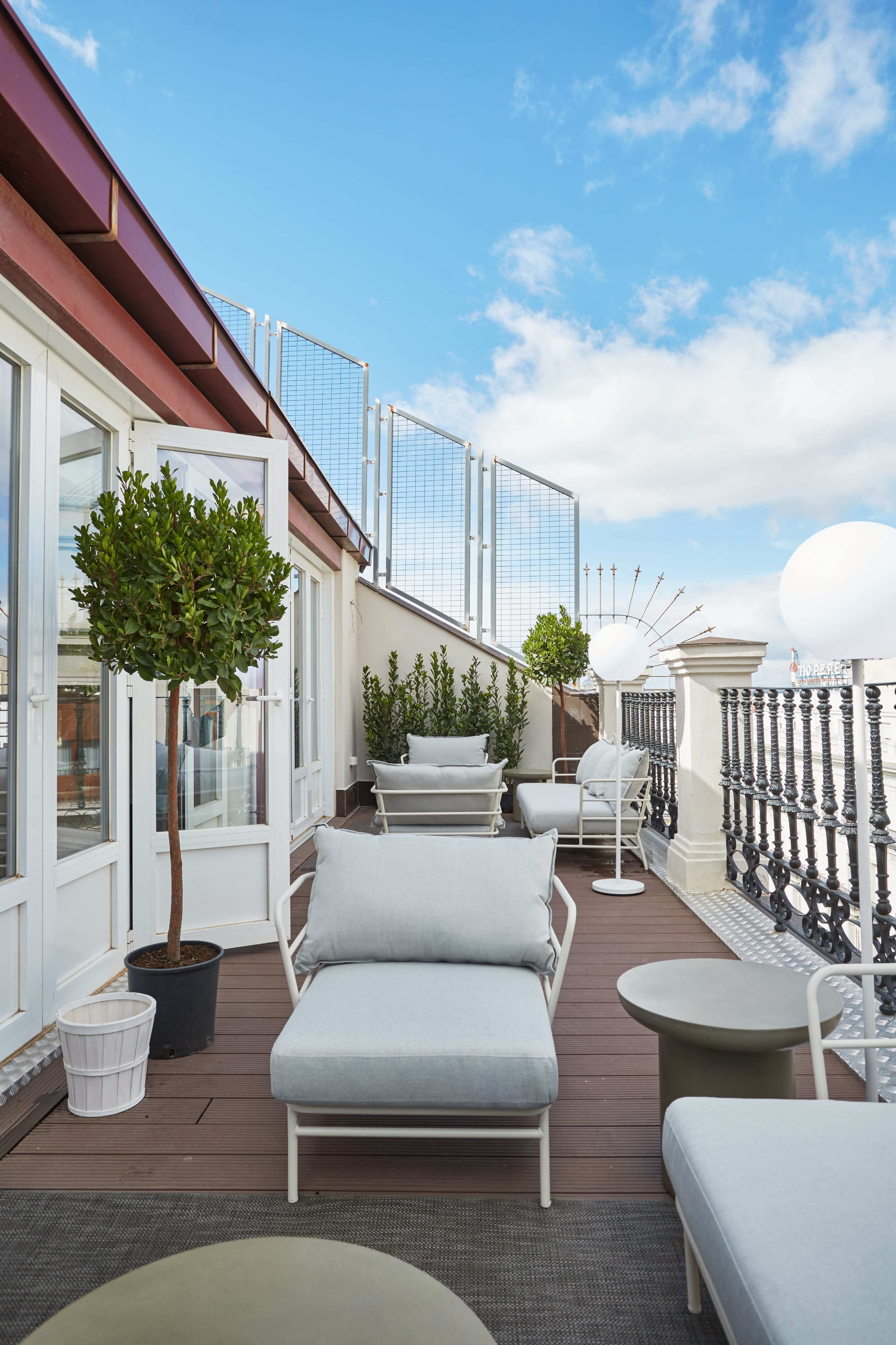 The image shows a rooftop terrace with modern outdoor seating, potted plants, and a clear blue sky.
