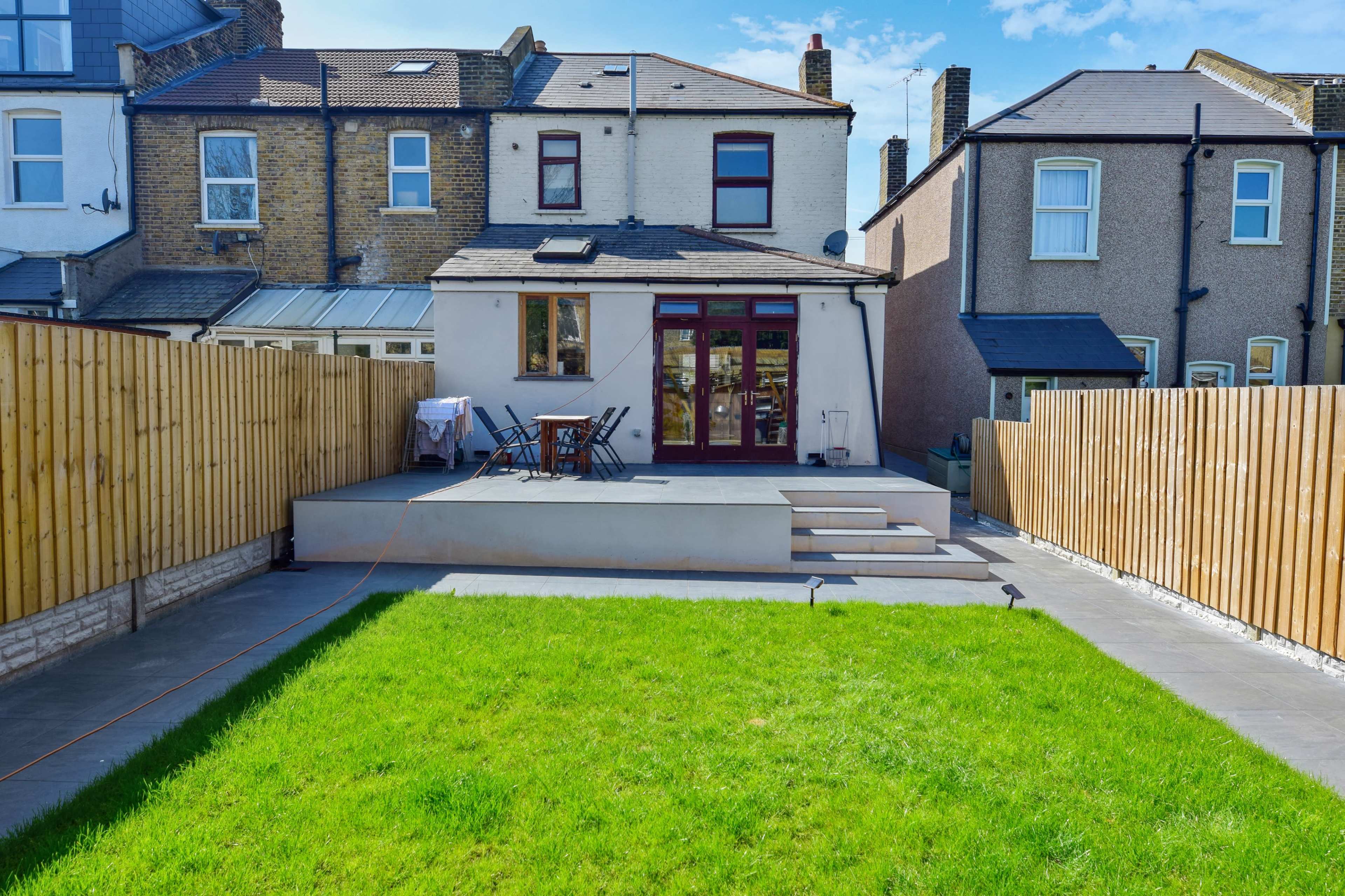A backyard with a lawn, a paved patio area, and a two-story house in the background, enclosed by wooden fences.