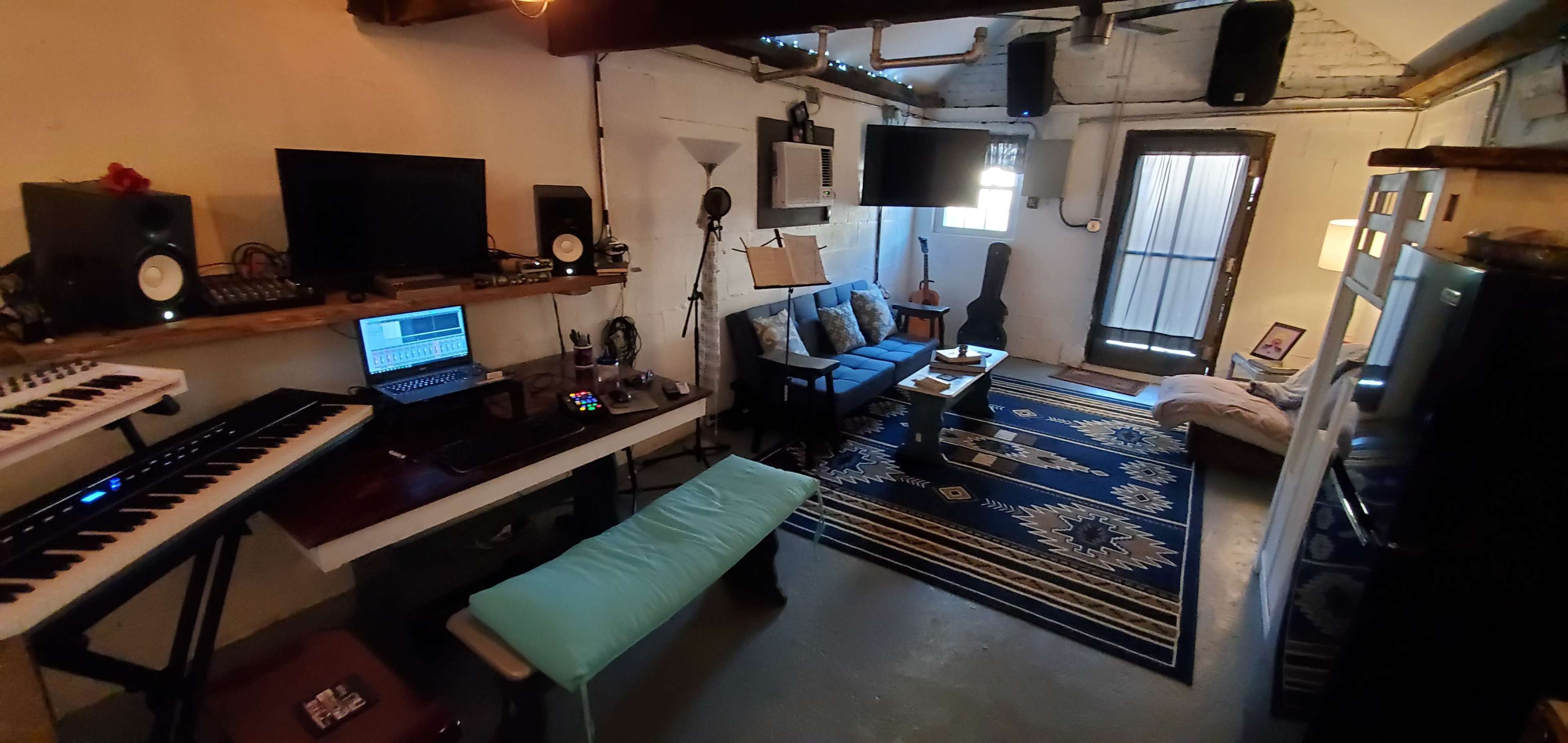 The image depicts a cozy studio space featuring a small seating area with a blue couch, a work desk with music equipment, and a door leading outside.