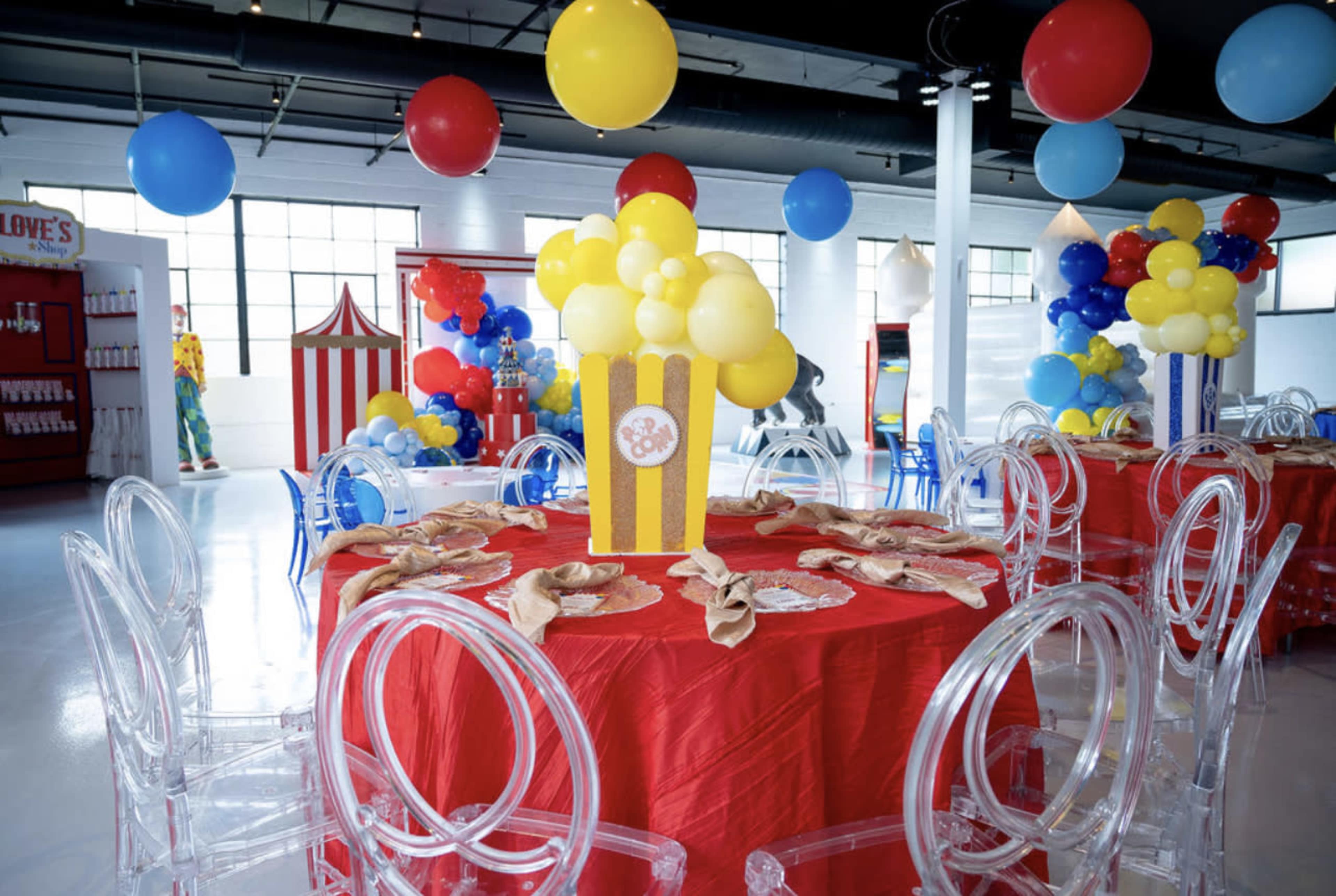 A vibrant party room features circular tables set with yellow and red decor, clear acrylic chairs, and colorful balloon arrangements.