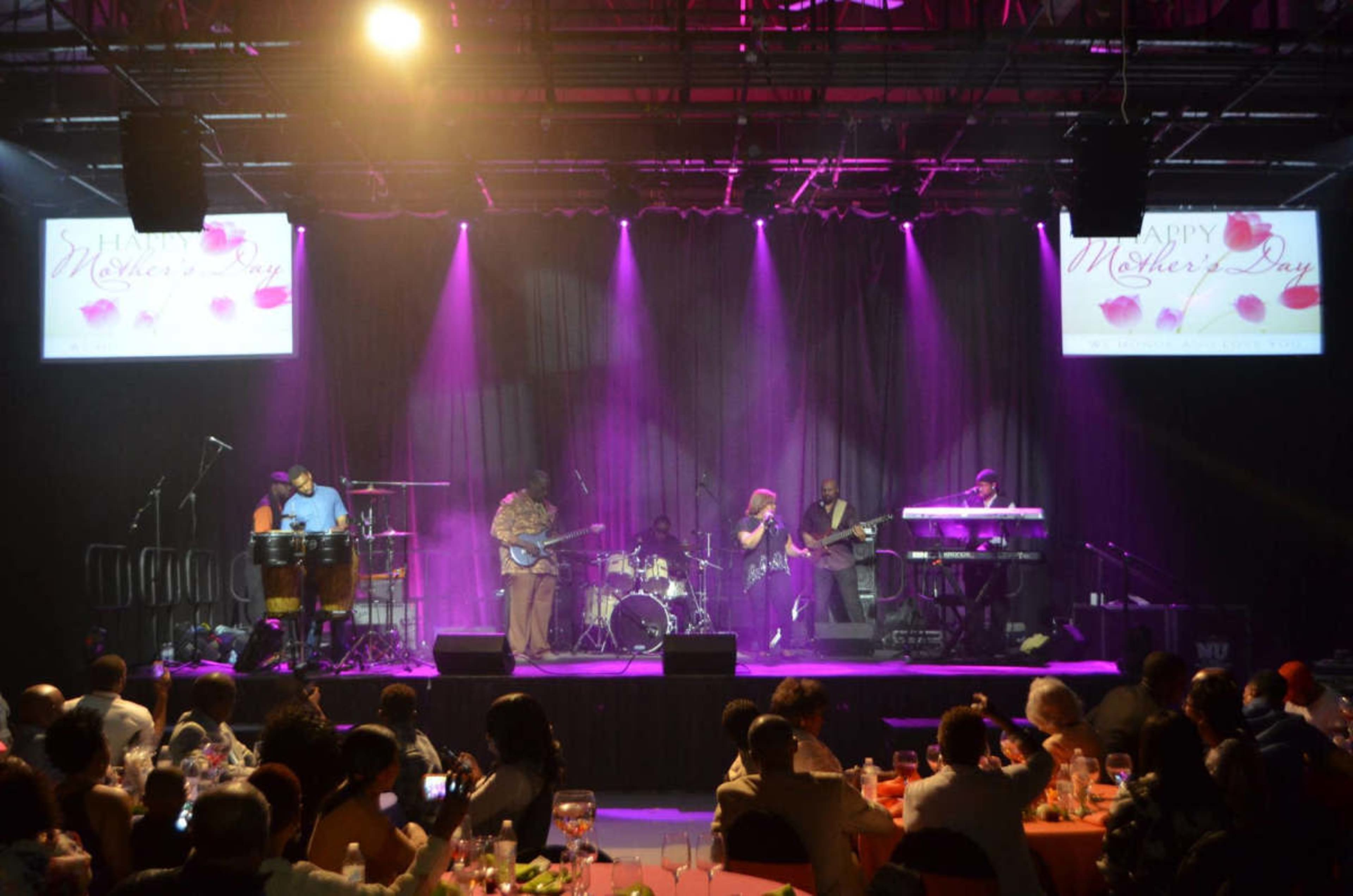 A musical band performs on stage in front of an audience during a Mother's Day event, with colorful lighting and decorations.