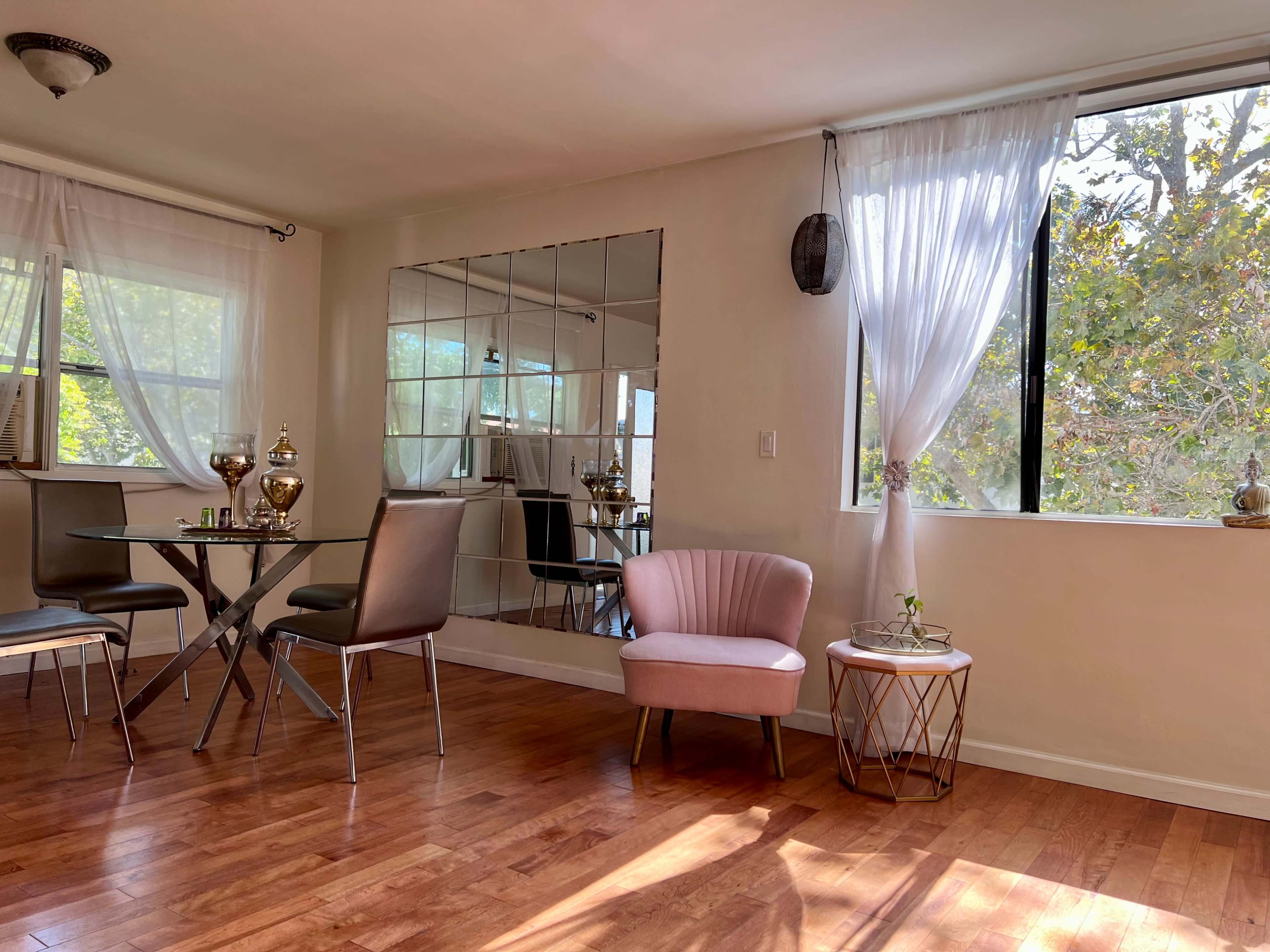 A bright dining area with a glass table, four chairs, a pink armchair, and a large mirror reflecting the window and natural light.
