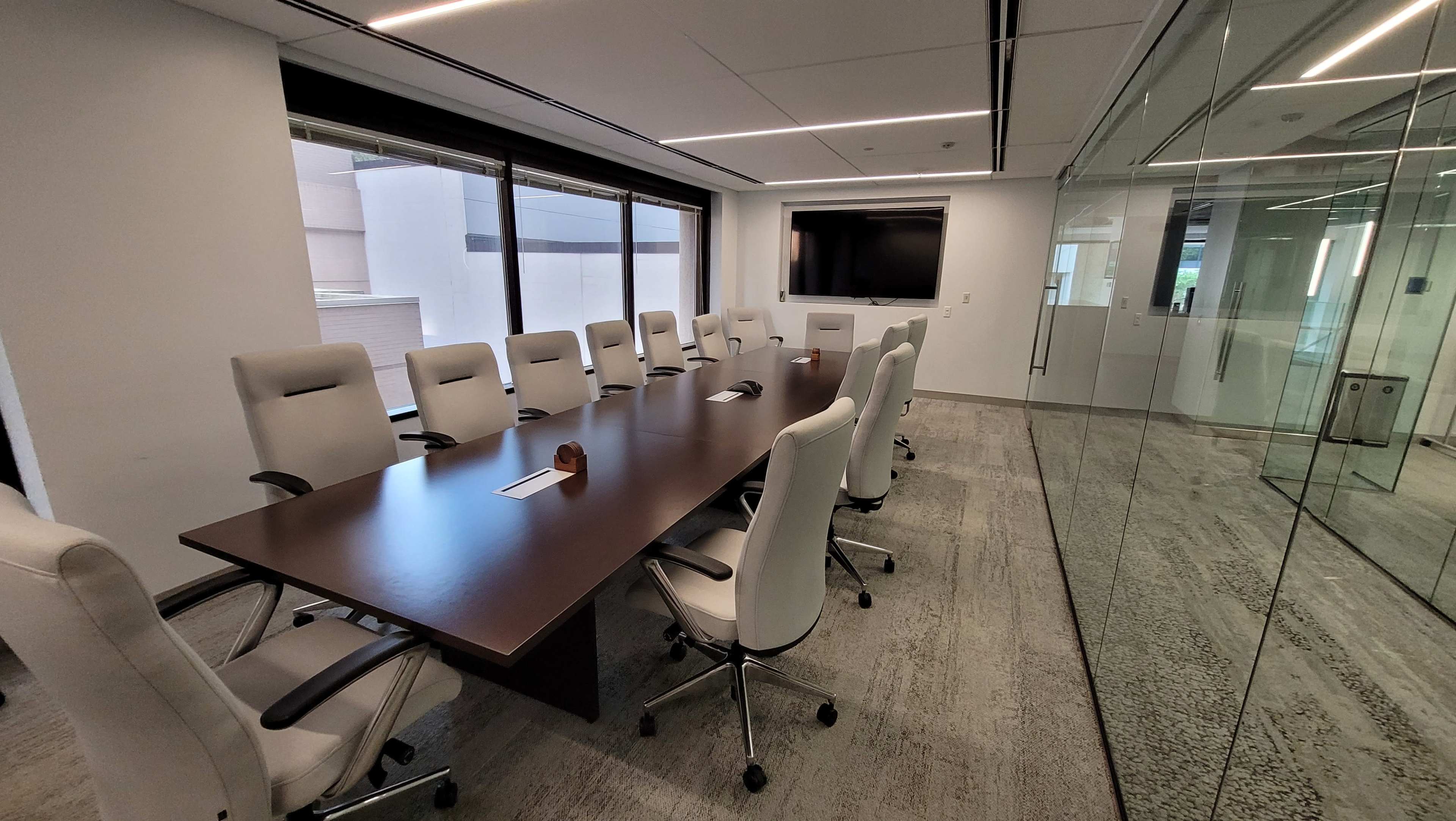 The image shows a modern conference room featuring a large rectangular table surrounded by ten white chairs, with a glass wall and a wall-mounted screen.