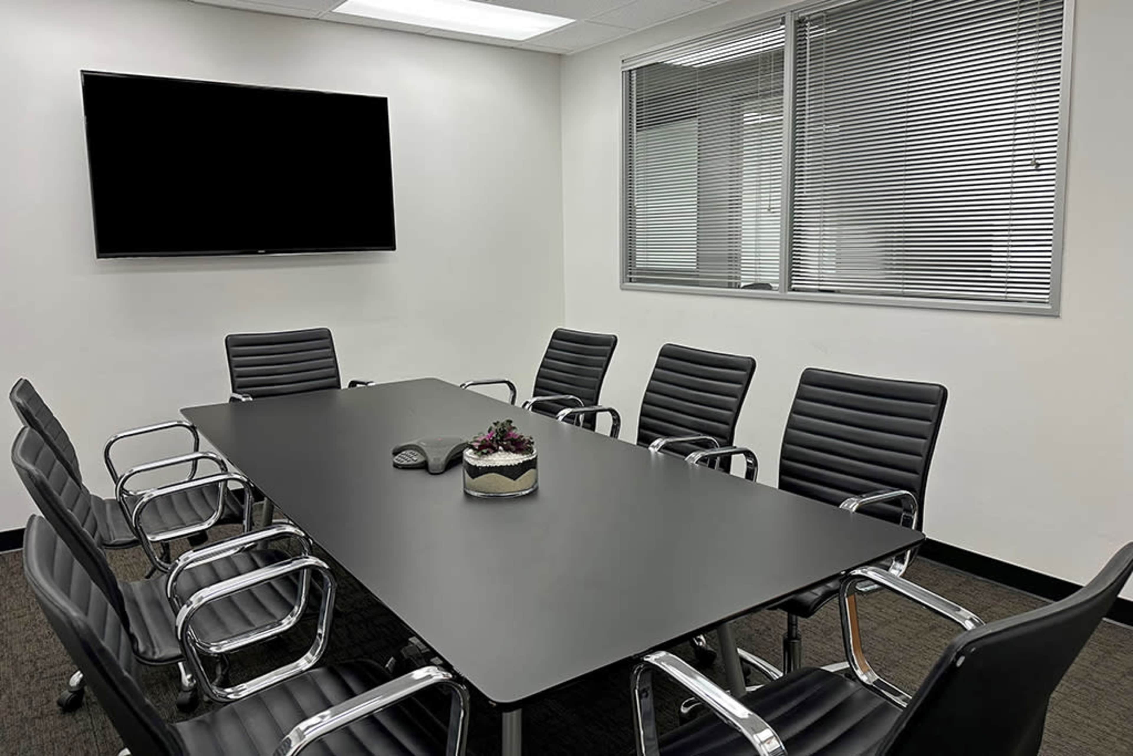 A modern conference room features a long black table surrounded by eight black leather chairs and a wall-mounted television.