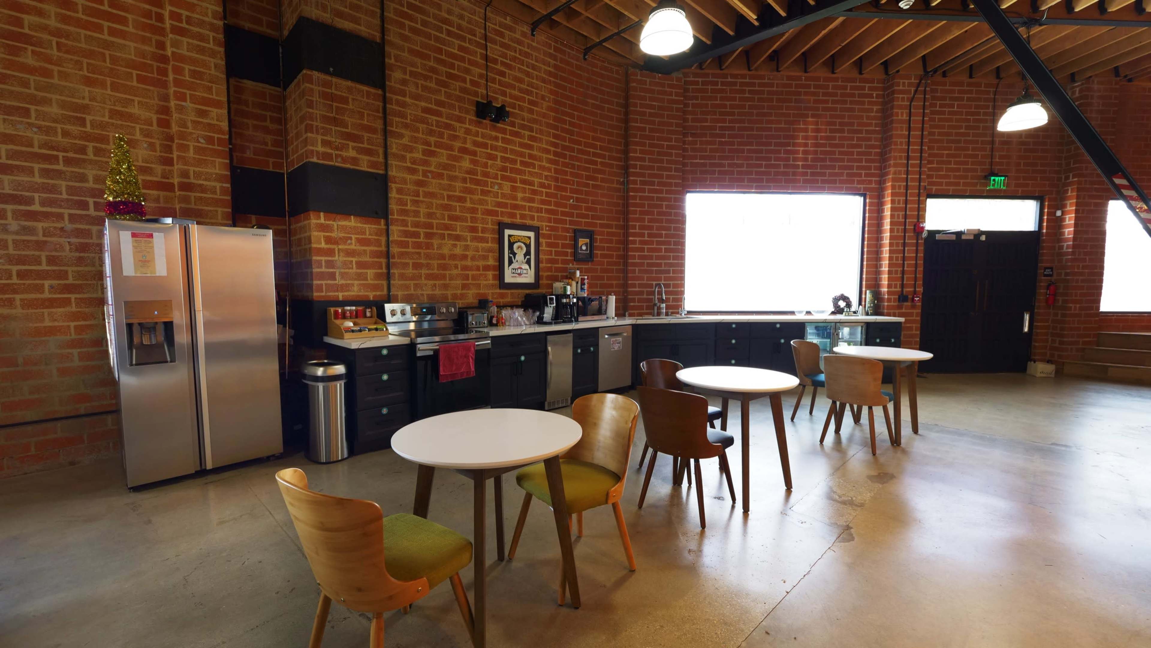 The image shows a modern kitchen space with brick walls, stainless steel appliances, and several round tables surrounded by wooden chairs.