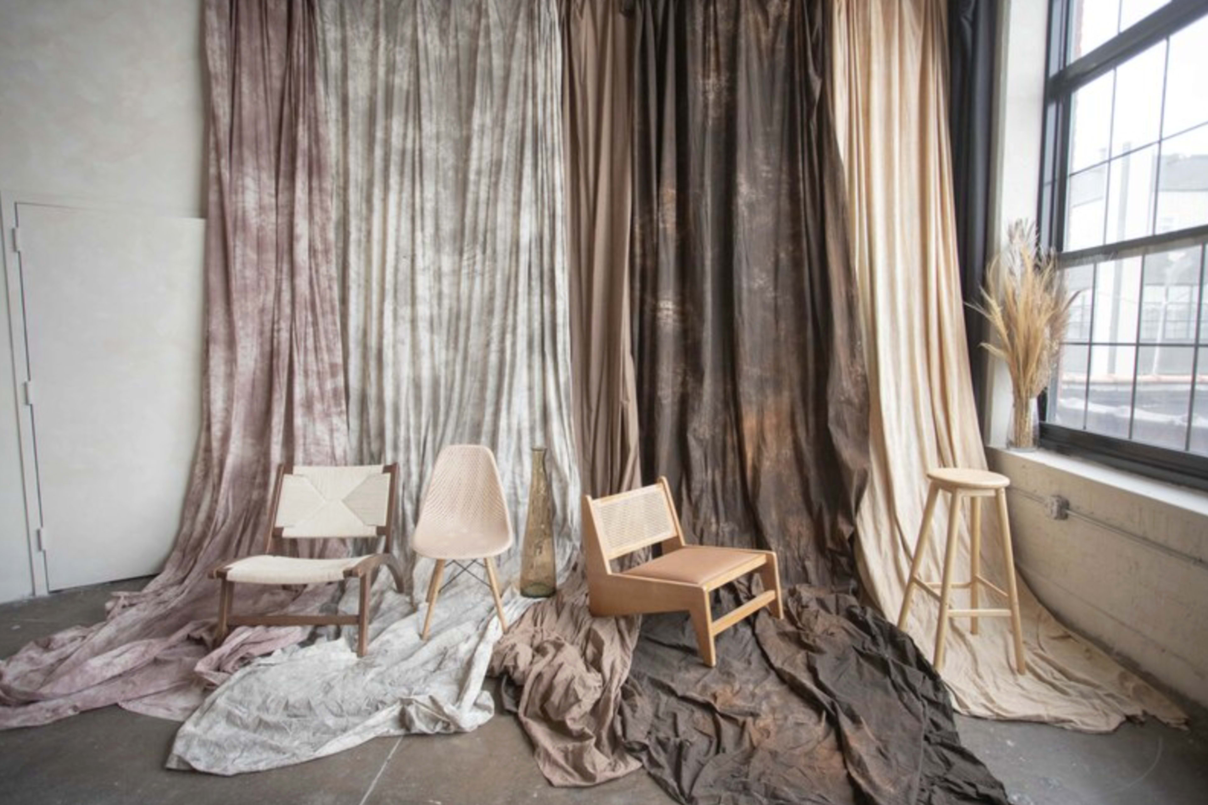 The image features a minimalist interior with a variety of draped fabrics in soft tones and four distinct chairs arranged in front of them.