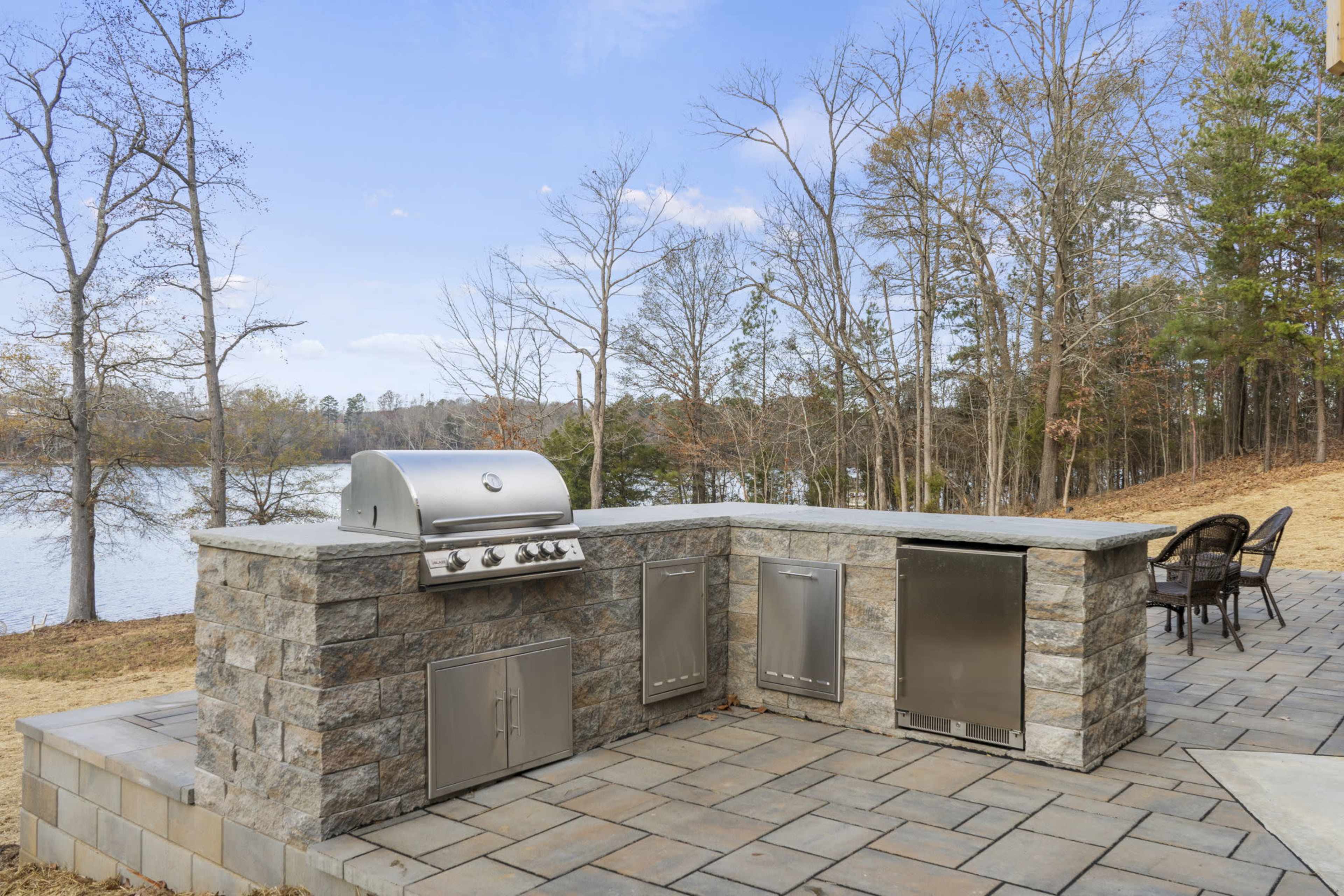The image shows an outdoor kitchen area with a stainless steel grill and storage cabinets set on a stone patio beside a lake.