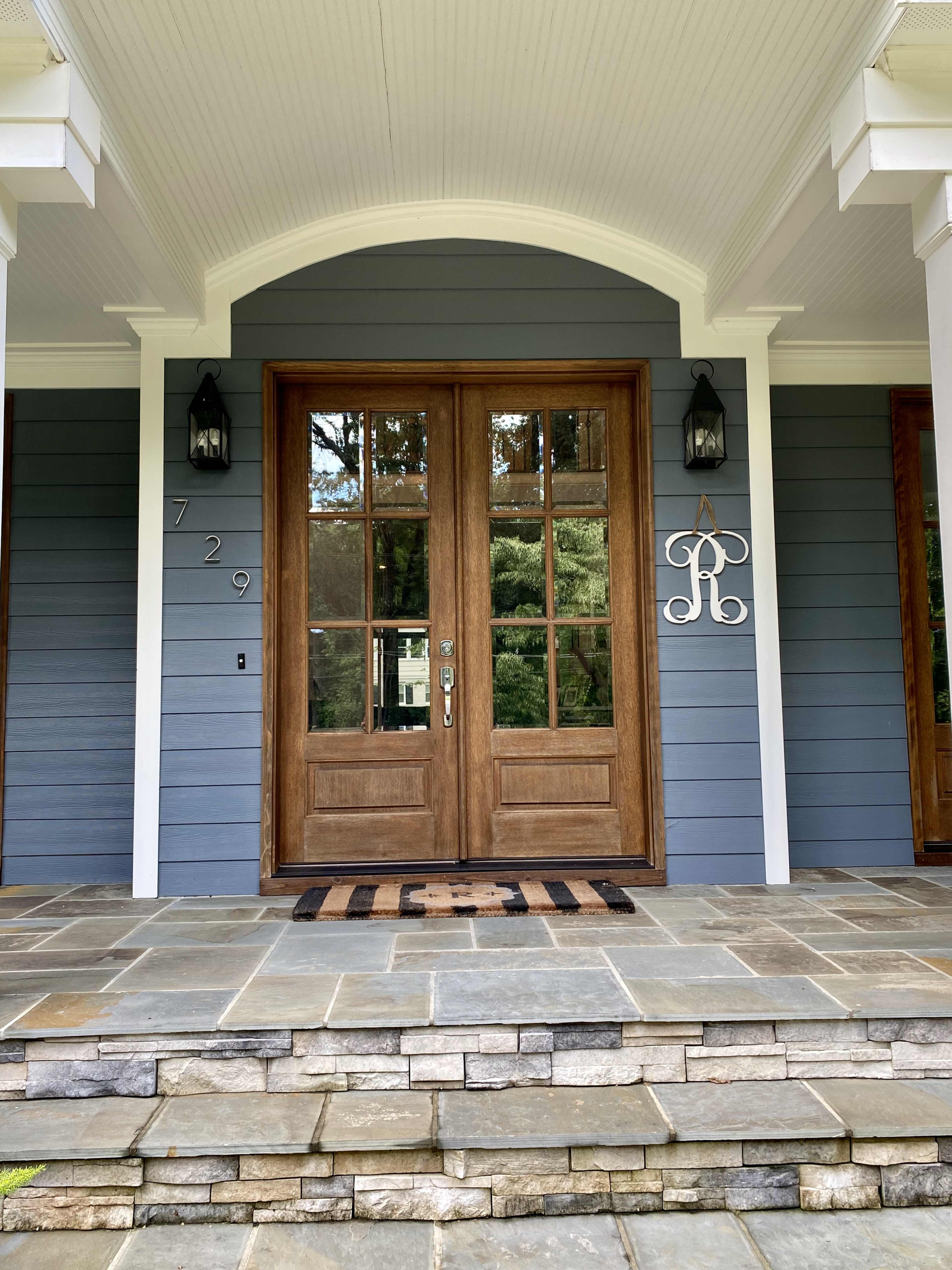 A wooden front door with glass panels, framed by blue siding and stone steps, featuring a welcome mat and decorative monogram.
