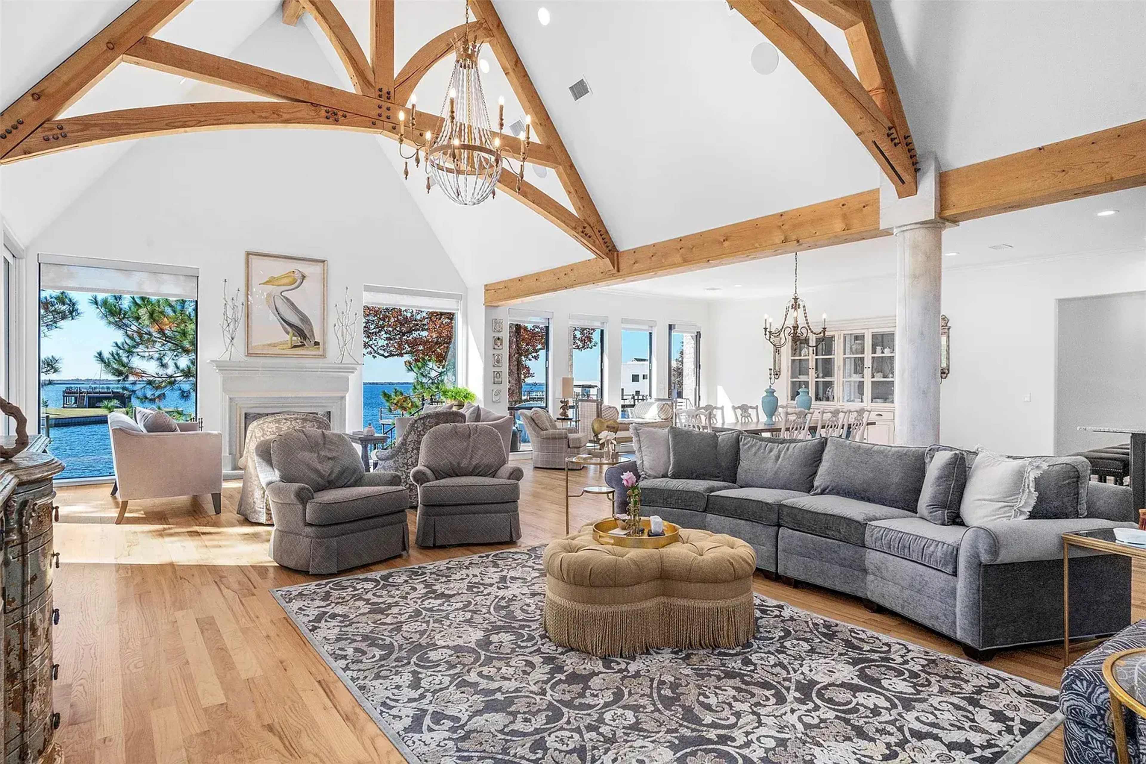 The spacious living room features high wooden beams, a decorative chandelier, a large sectional sofa, and views of the outdoor scenery through expansive windows.