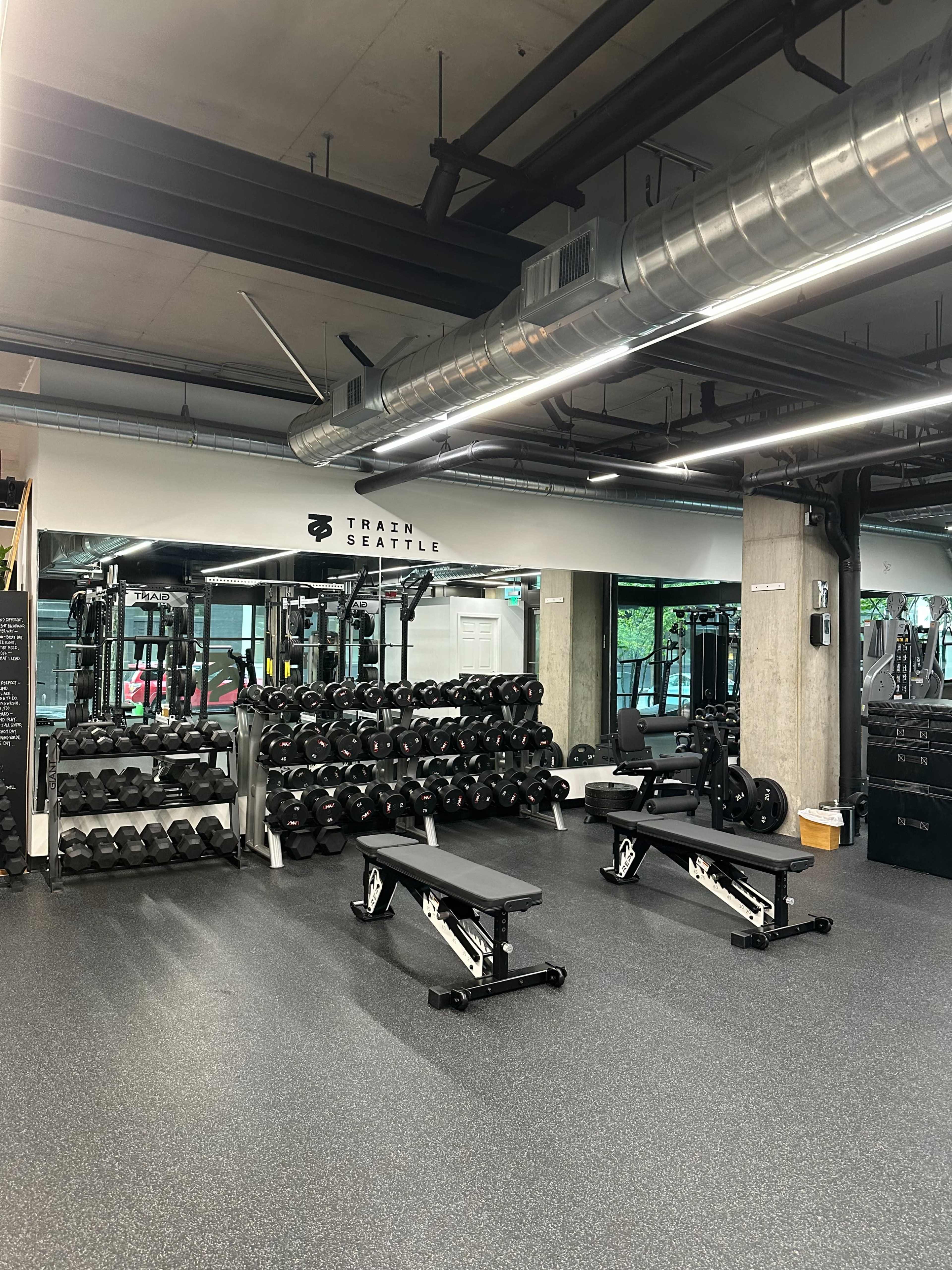 The image shows a modern gym interior featuring a weightlifting area with dumbbells, benches, and various exercise equipment.