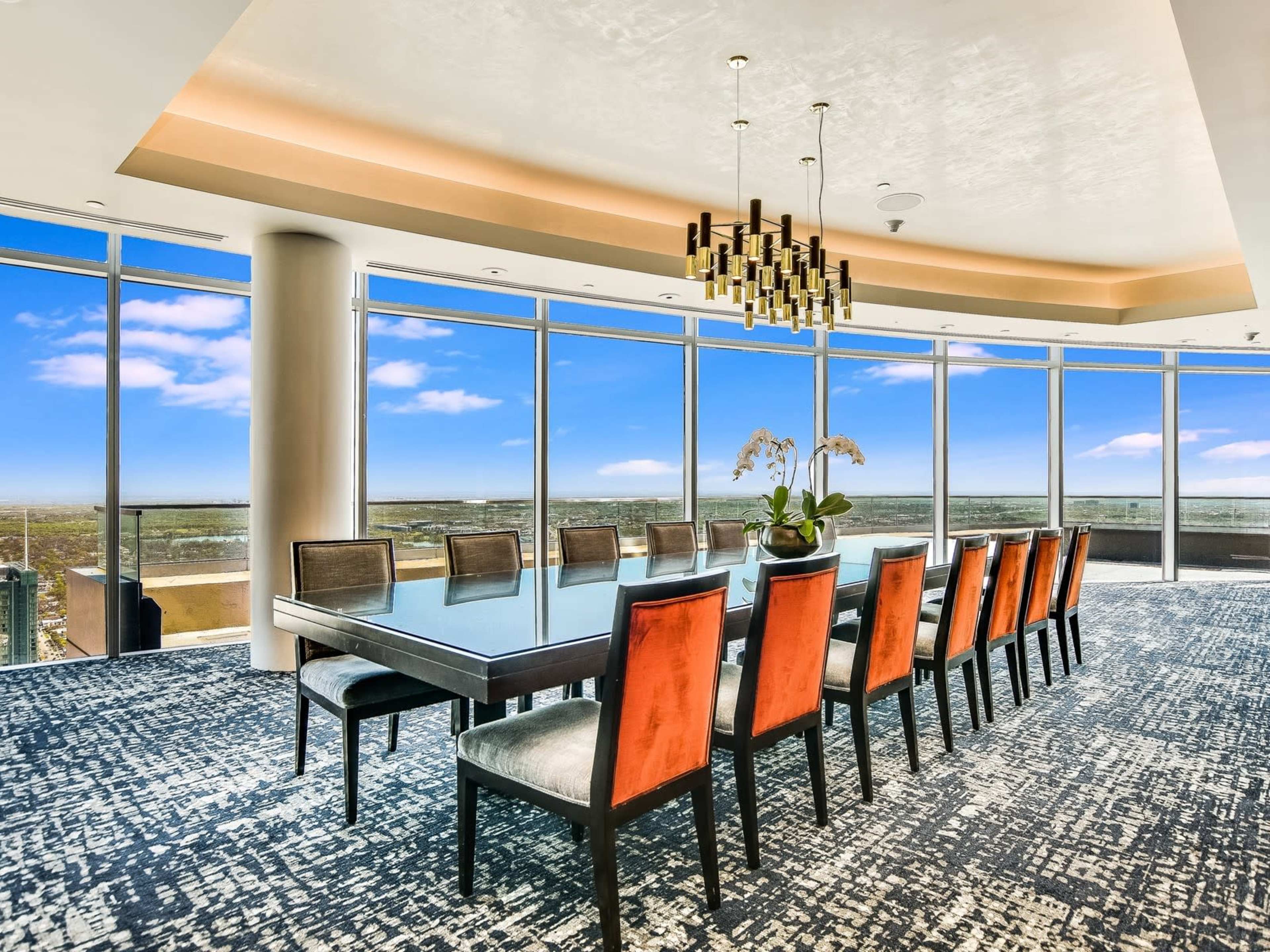 A large dining table is set with twelve chairs in a modern, brightly lit room featuring floor-to-ceiling windows that offer a view of the landscape outside.