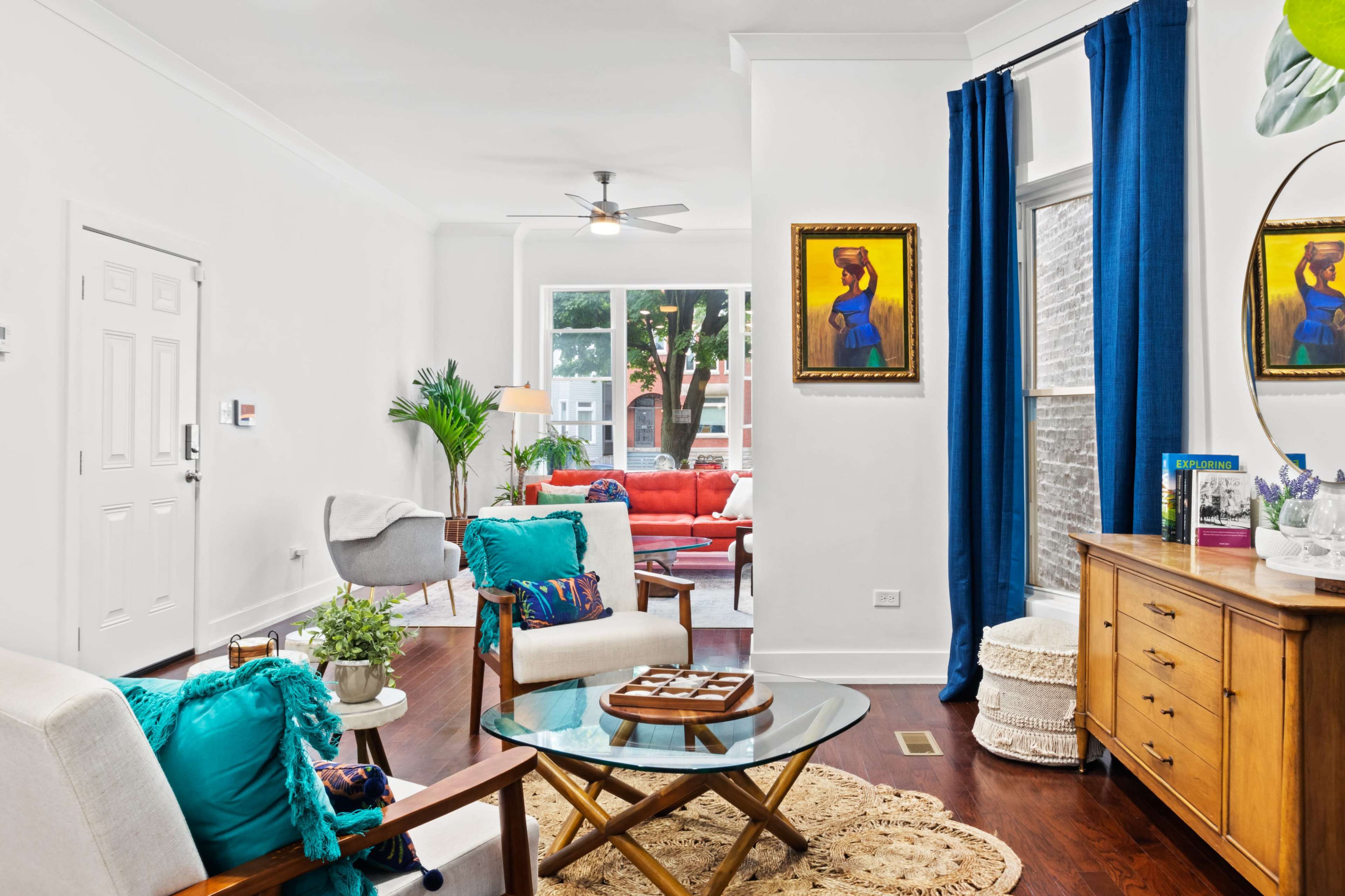 A bright living room featuring a mix of colorful furniture, potted plants, and decorative artwork against white walls.