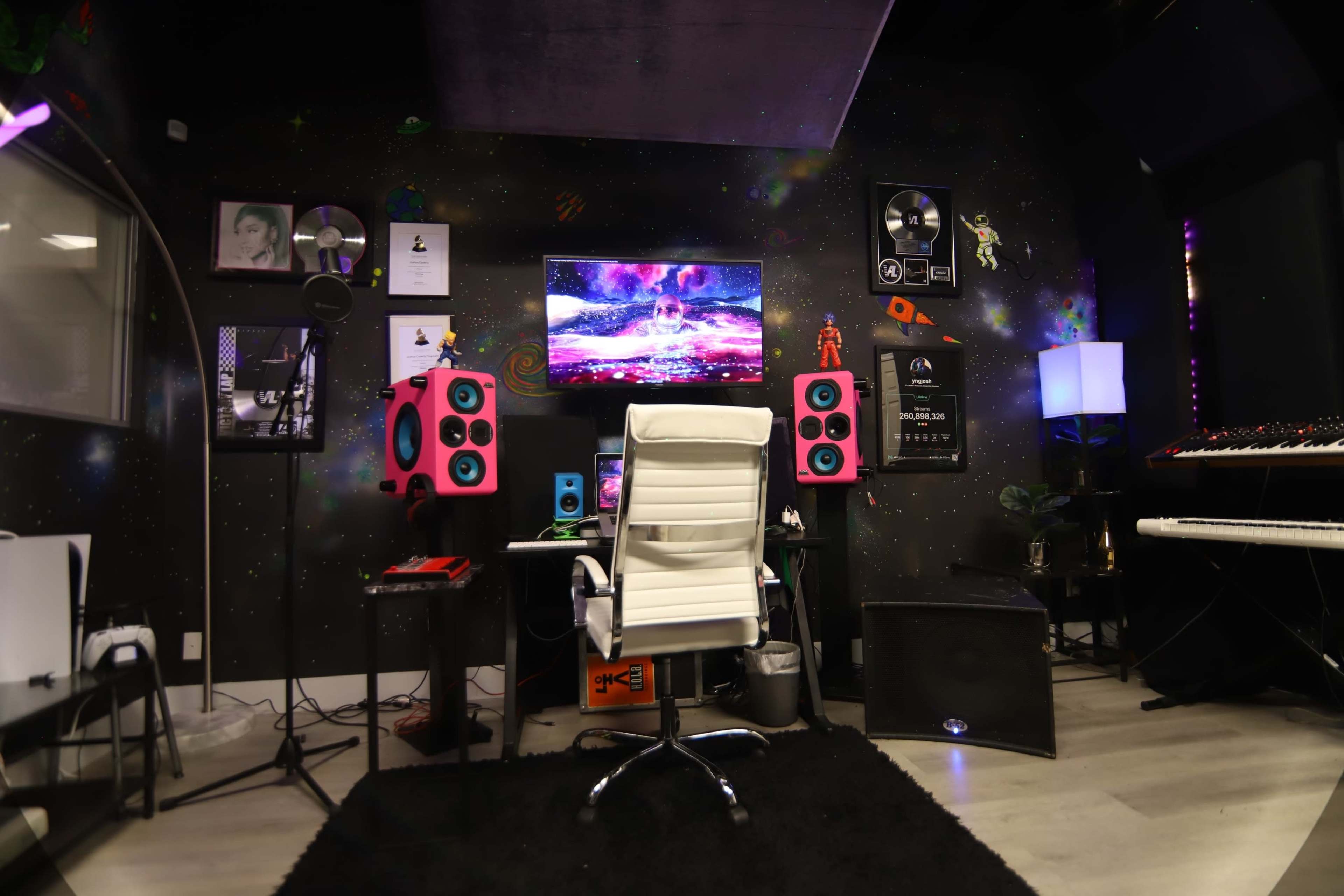 A music studio featuring a white office chair in front of a television, pink speakers on either side, a black rug on the floor, and various musical equipment and decorations on the walls.