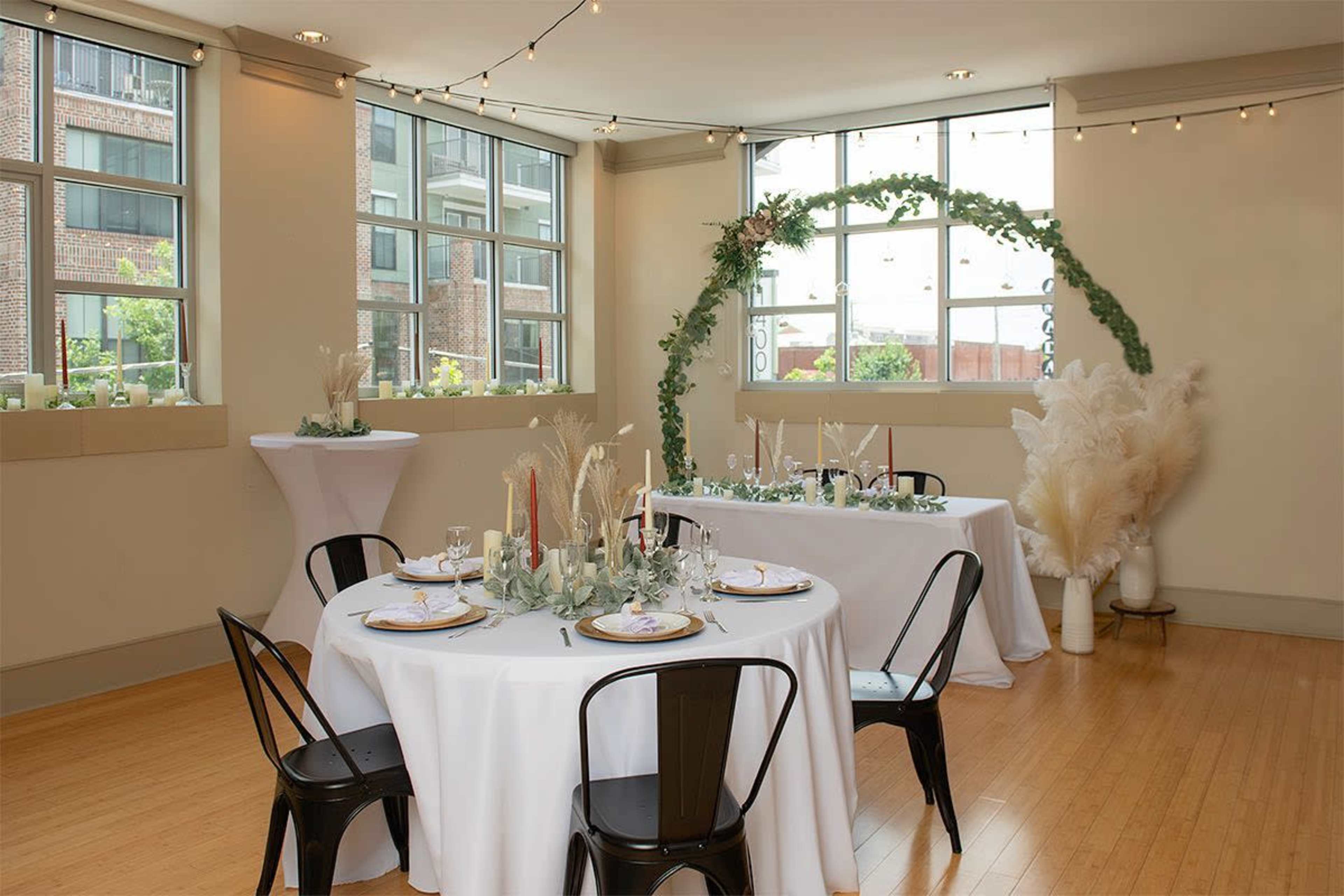 A decorated event space with two tables set for dining, featuring candles, floral arrangements, and an arch made of greenery near large windows.