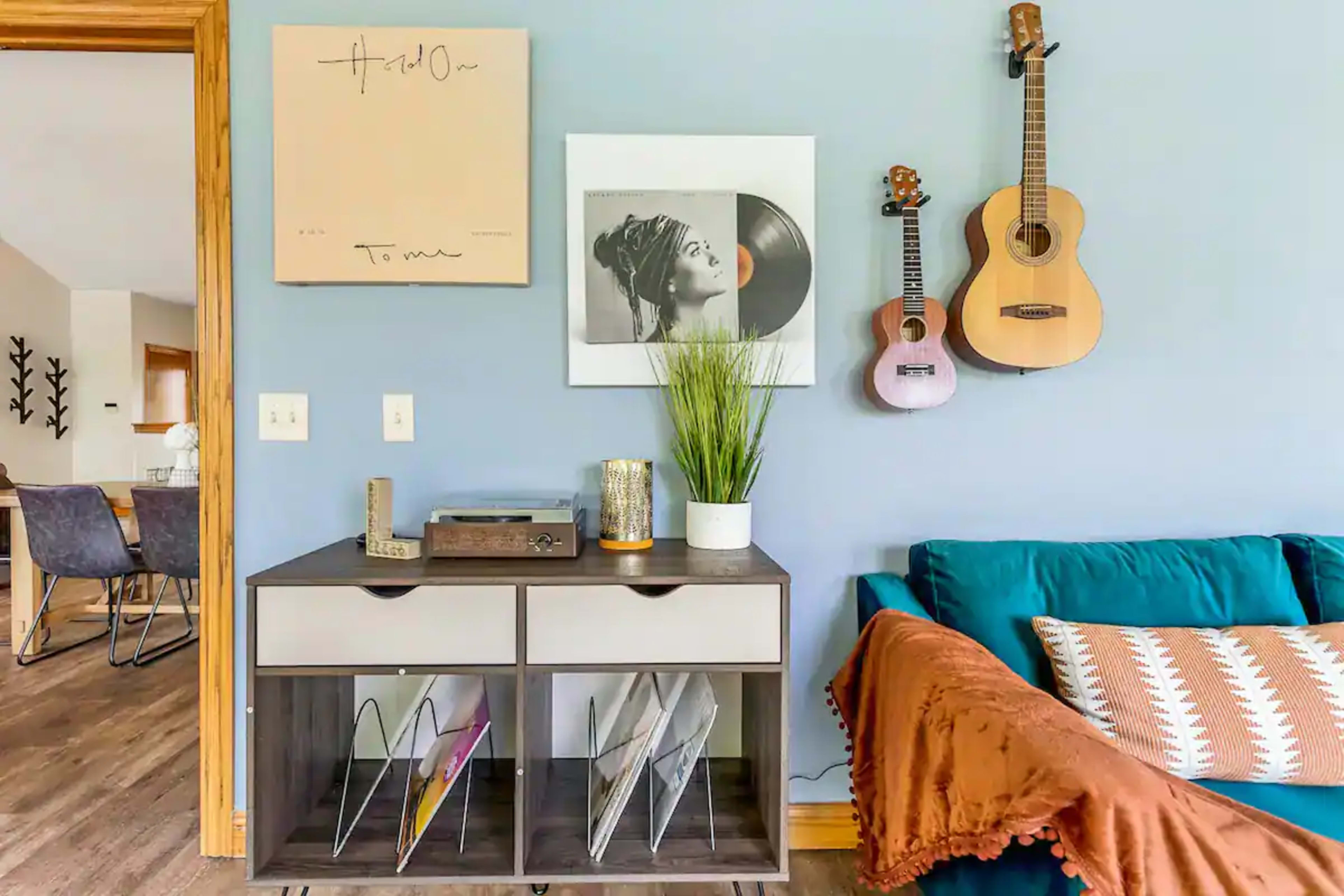 A console table with magazines, a record player, and decorative items is positioned against a light blue wall adorned with framed art and two guitars.