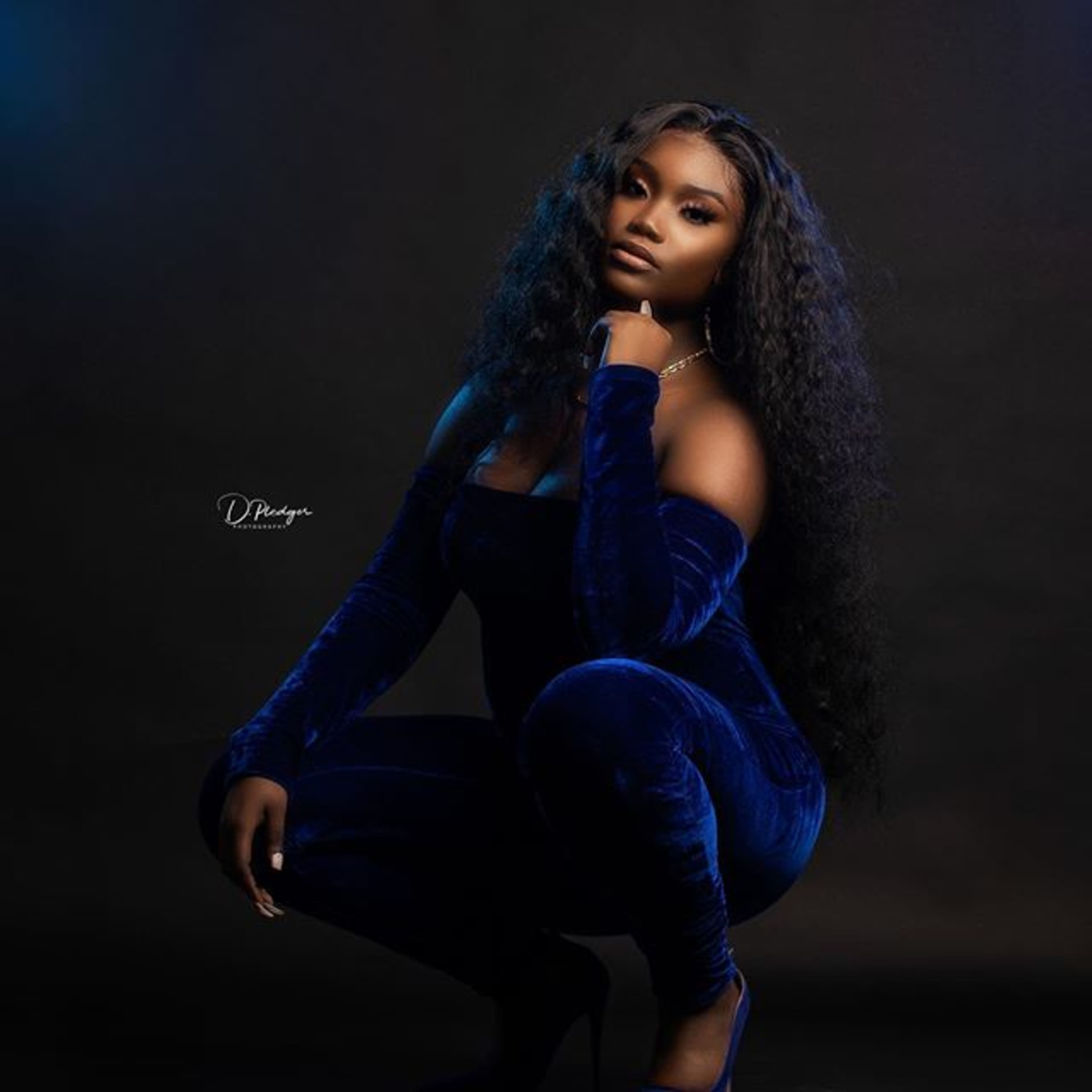 A woman in a dark blue, off-shoulder jumpsuit poses in a crouched position against a black background.