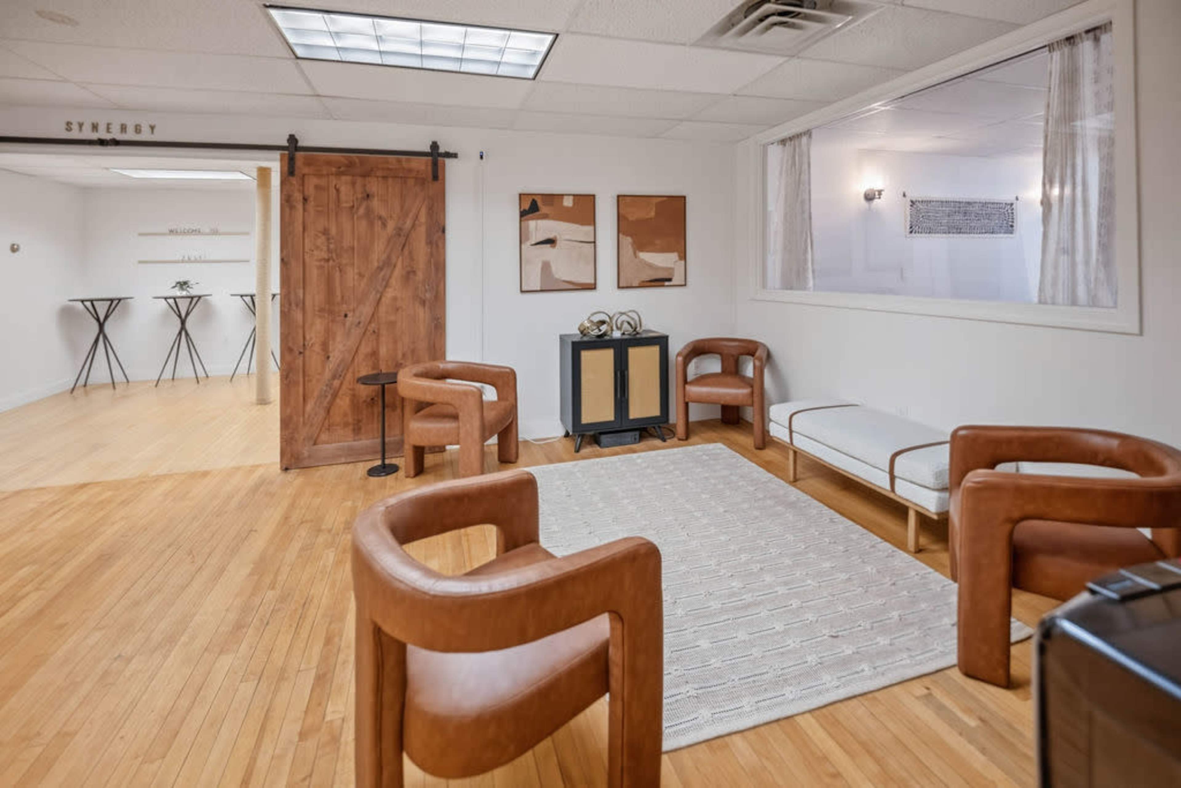 The image shows a spacious, modern room with wooden flooring, featuring a simple seating arrangement with brown chairs, a white couch, and a door with a wooden sliding design.