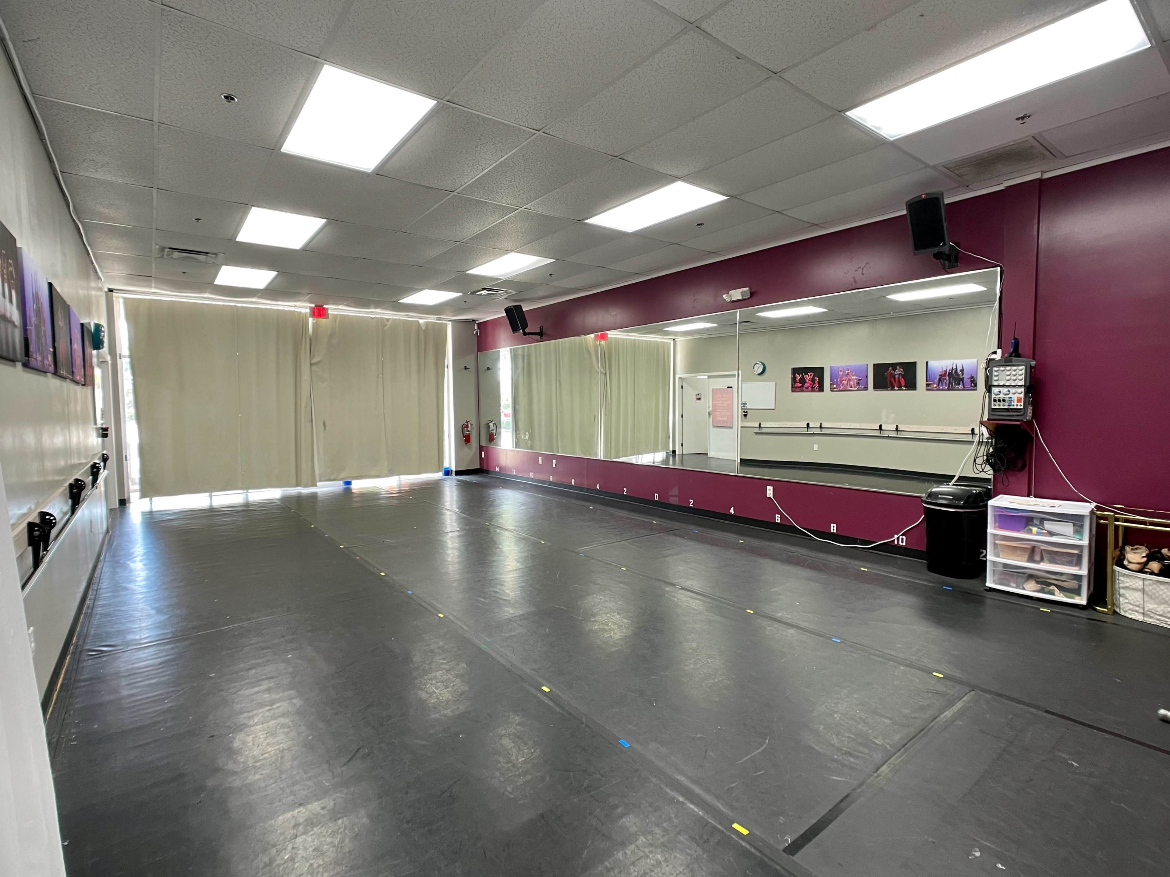 A dance studio features a large mirrored wall, a smooth black floor, and natural light coming through curtains at one end.