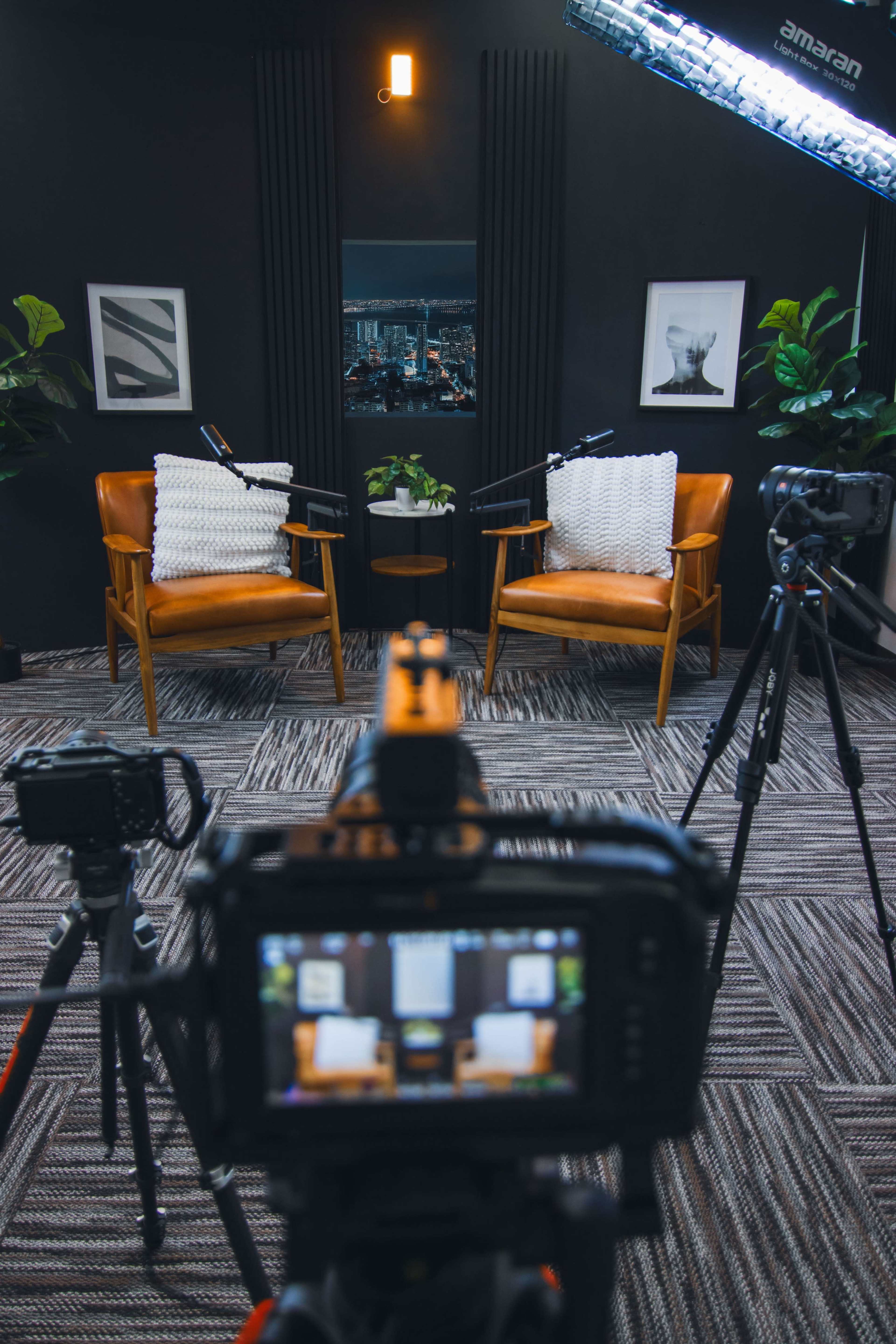 The image shows a well-lit interview setup featuring two chairs with decorative pillows, a small table, and cameras positioned to capture the scene in a studio environment.
