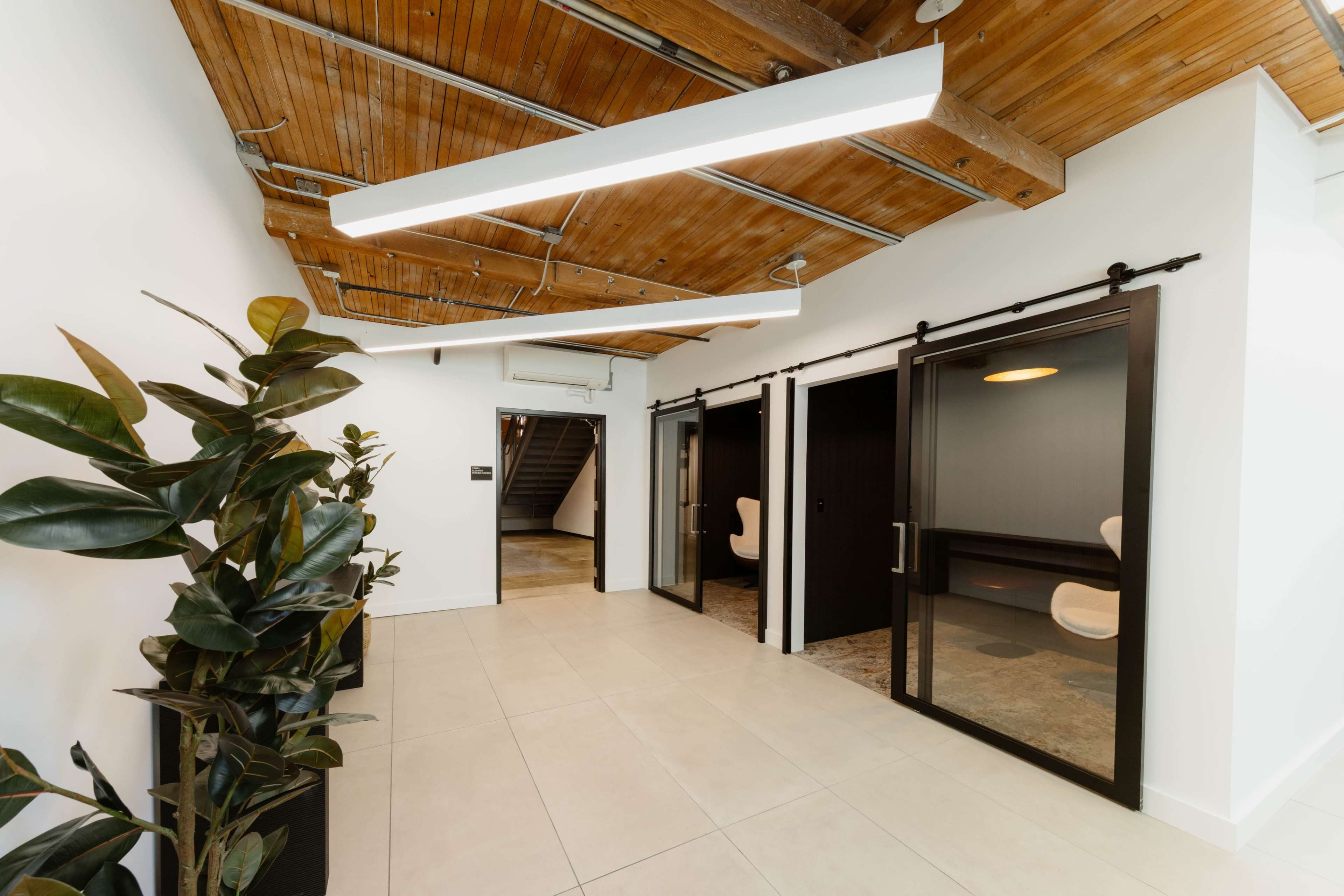 The image shows a modern hallway with wooden beams on the ceiling, potted plants on one side, and glass-walled meeting rooms opposite.