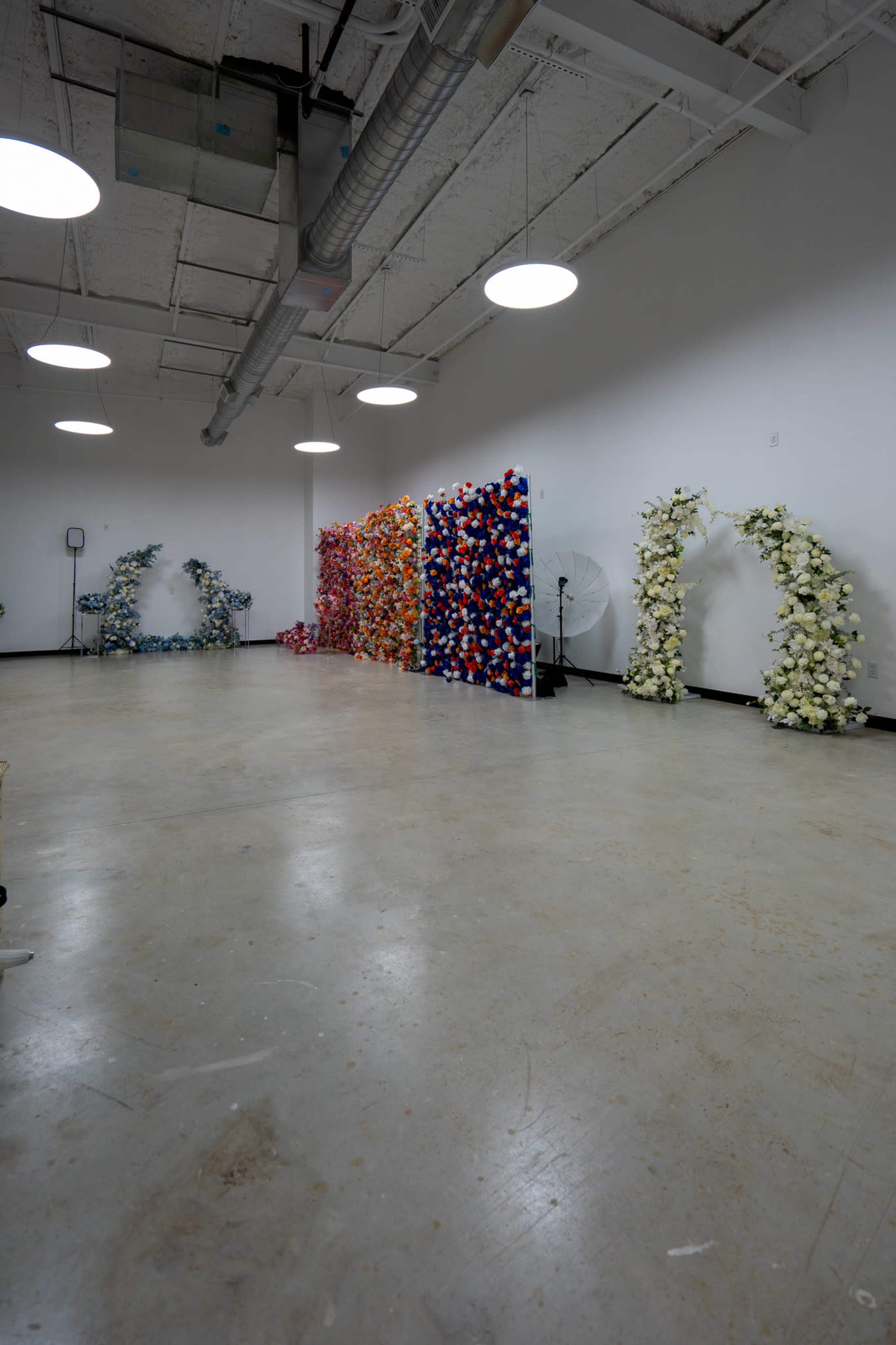 A spacious room with a concrete floor features a series of floral arrangements in various colors along one wall, with decorative arches on either end.