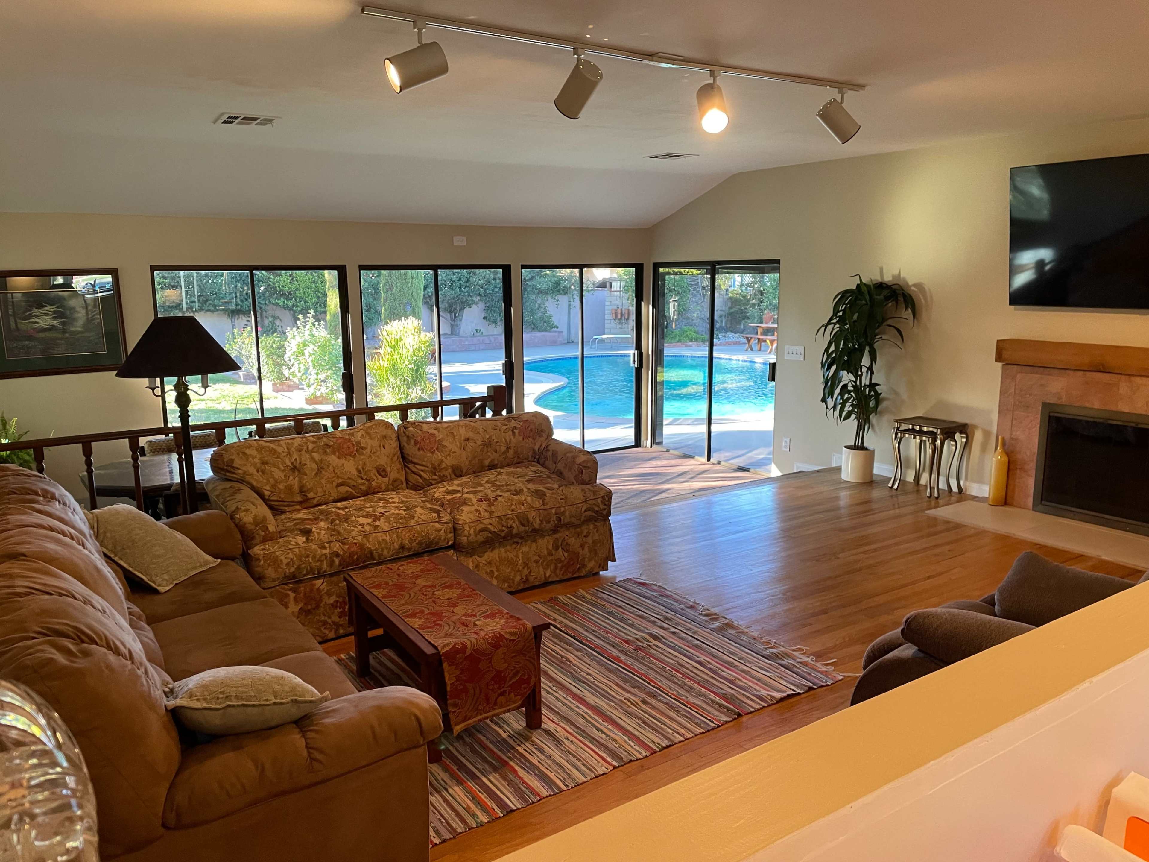 The image shows a living room with a large sofa, coffee table, and a view of a pool area through sliding glass doors.