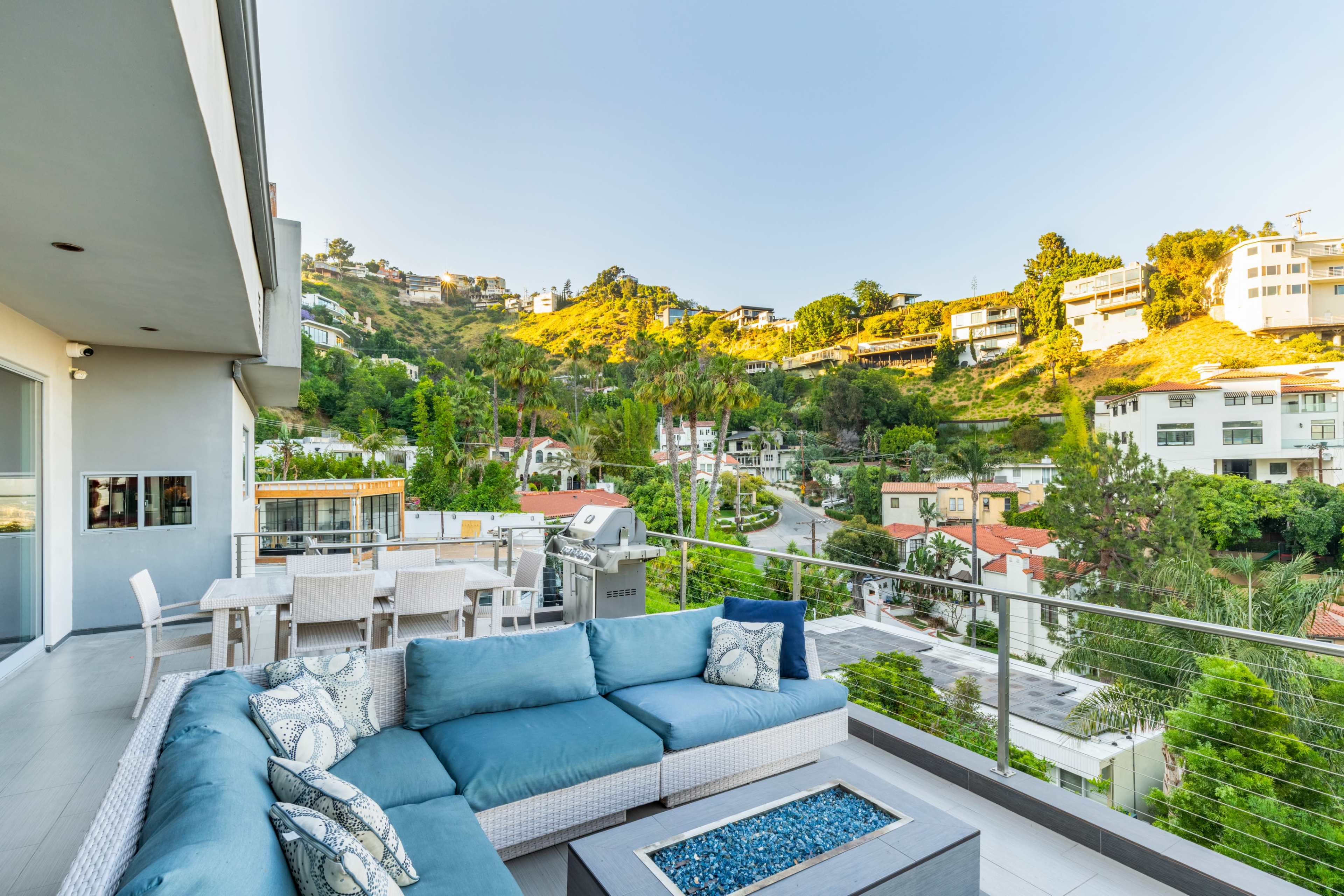 A spacious balcony features a blue sectional sofa, a fire pit, and a view of hillside homes surrounded by trees.