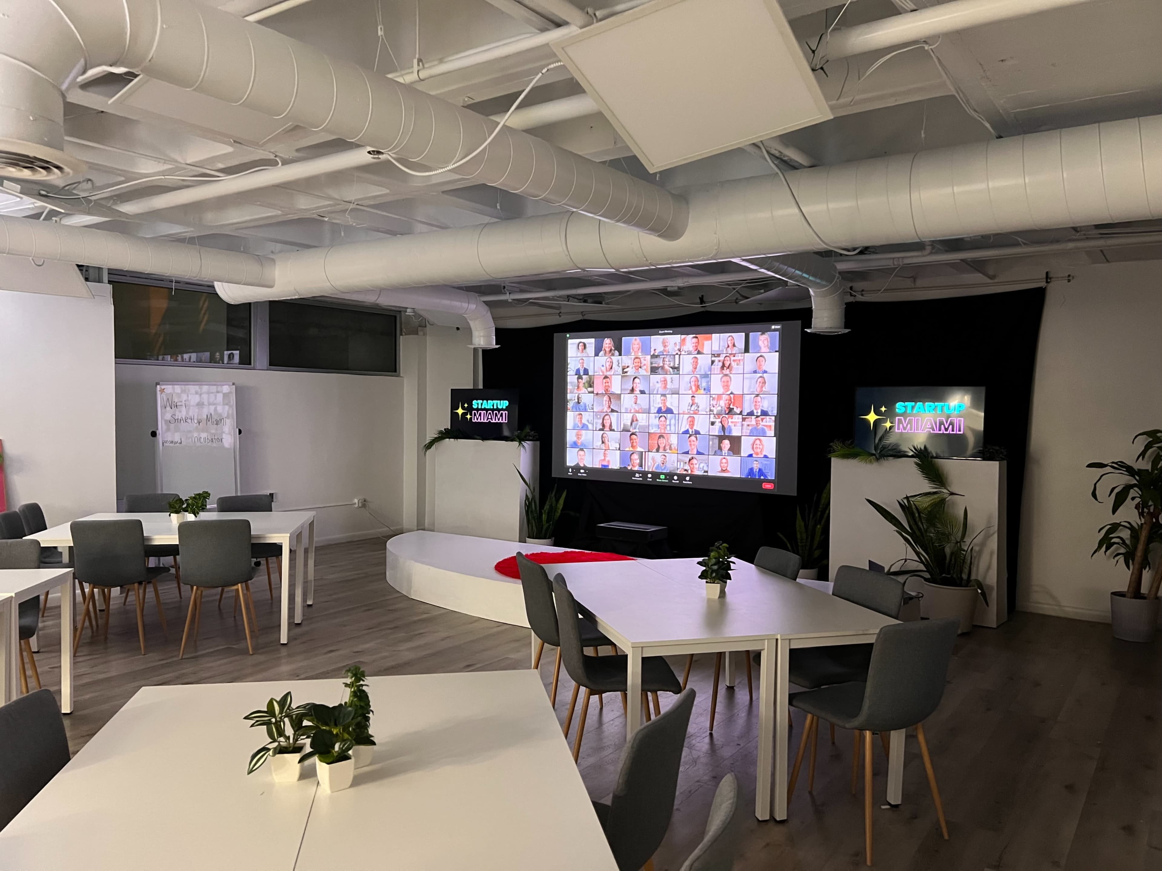 A modern meeting room features several tables, chairs, and a large screen displaying multiple participants in a virtual call, with greenery present throughout the space.